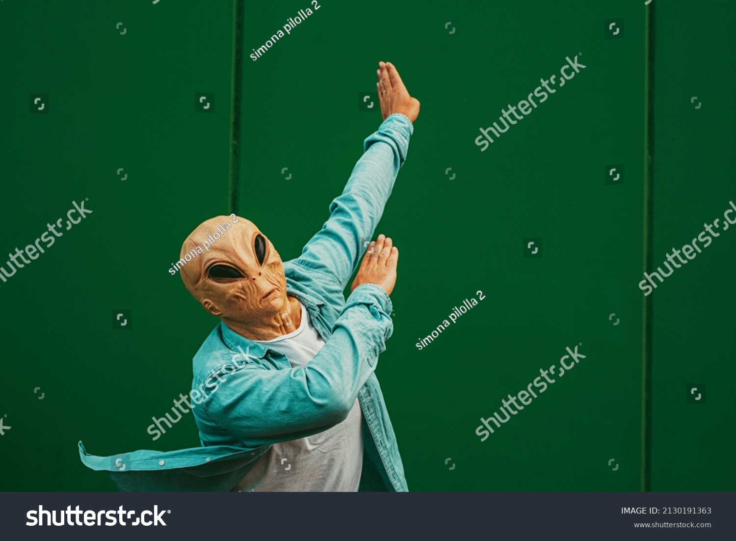 Alien doing dab posture and gesture against a green wall background. Extraterrestrial with human clothes. Concept of victory and satisfaction. Happiness and freedom immigration concept #2130191363