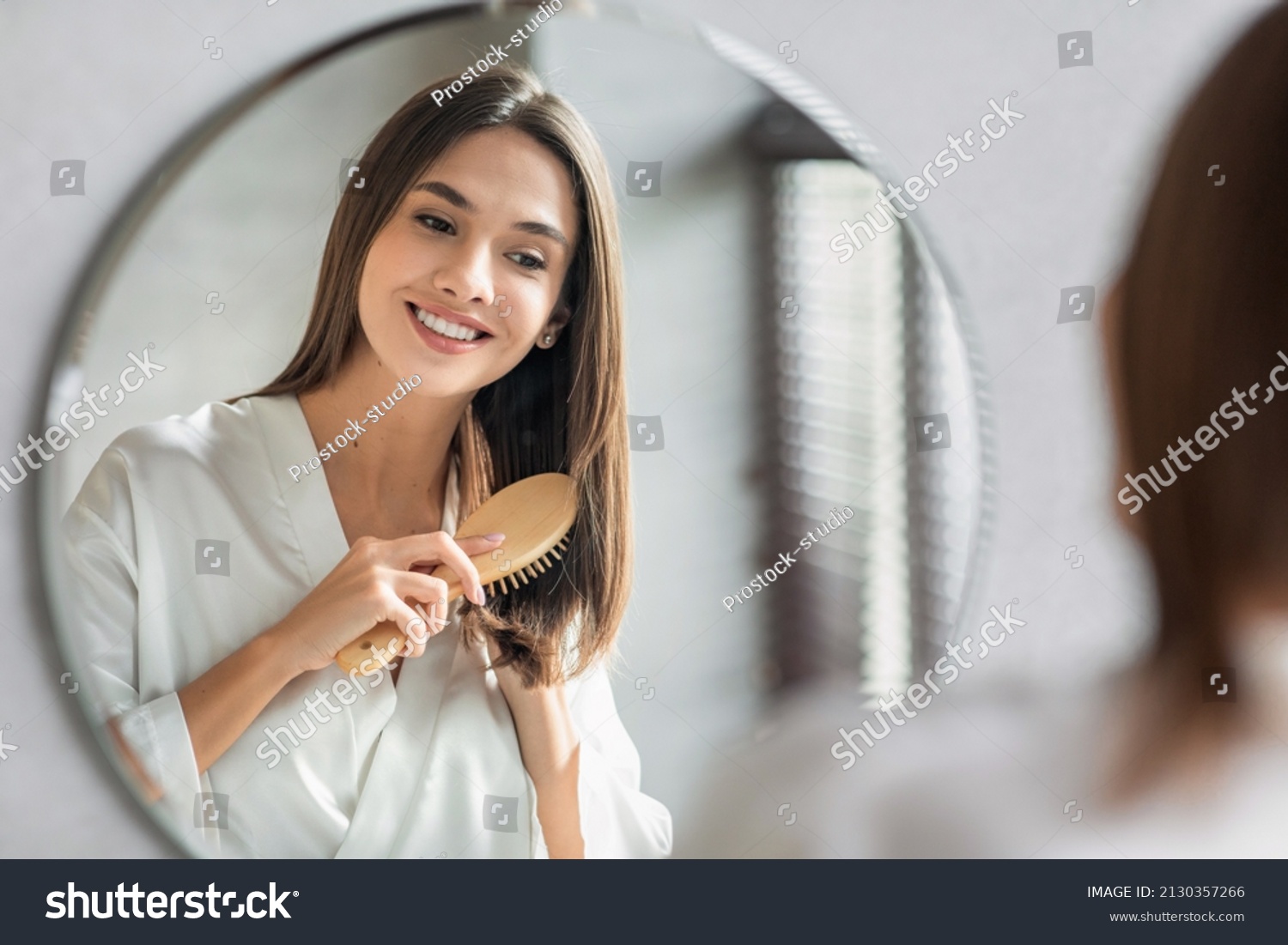 Beauty Routine. Pretty Woman Combing Her Beautiful Hair With Brush While Standing Near Mirror In Bathroom, Attractive Young Lady Looking To Her Reflection And Smiling, Selective Focus With Free Space #2130357266