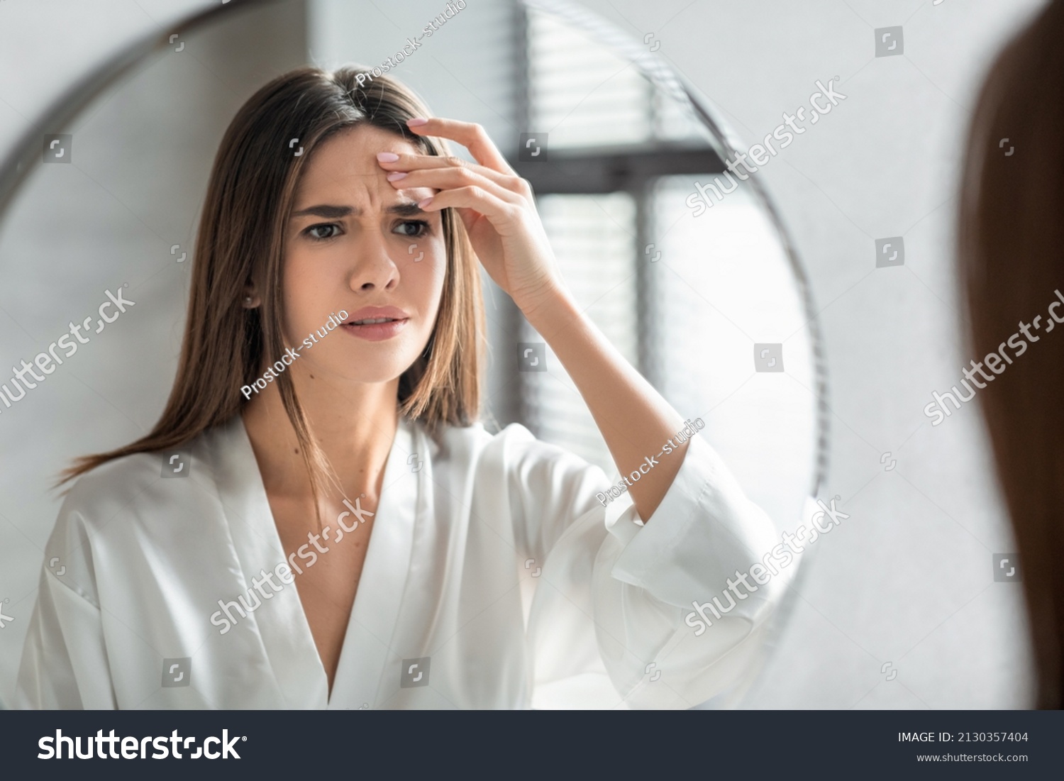 Unhappy Young Woman Looking In Mirror And Touching Wrinkles On Her Face, Attractive Millennial Female Standing In Bathroom And Examining Skin Fine Lines, Selective Focus On Reflection, Closeup #2130357404