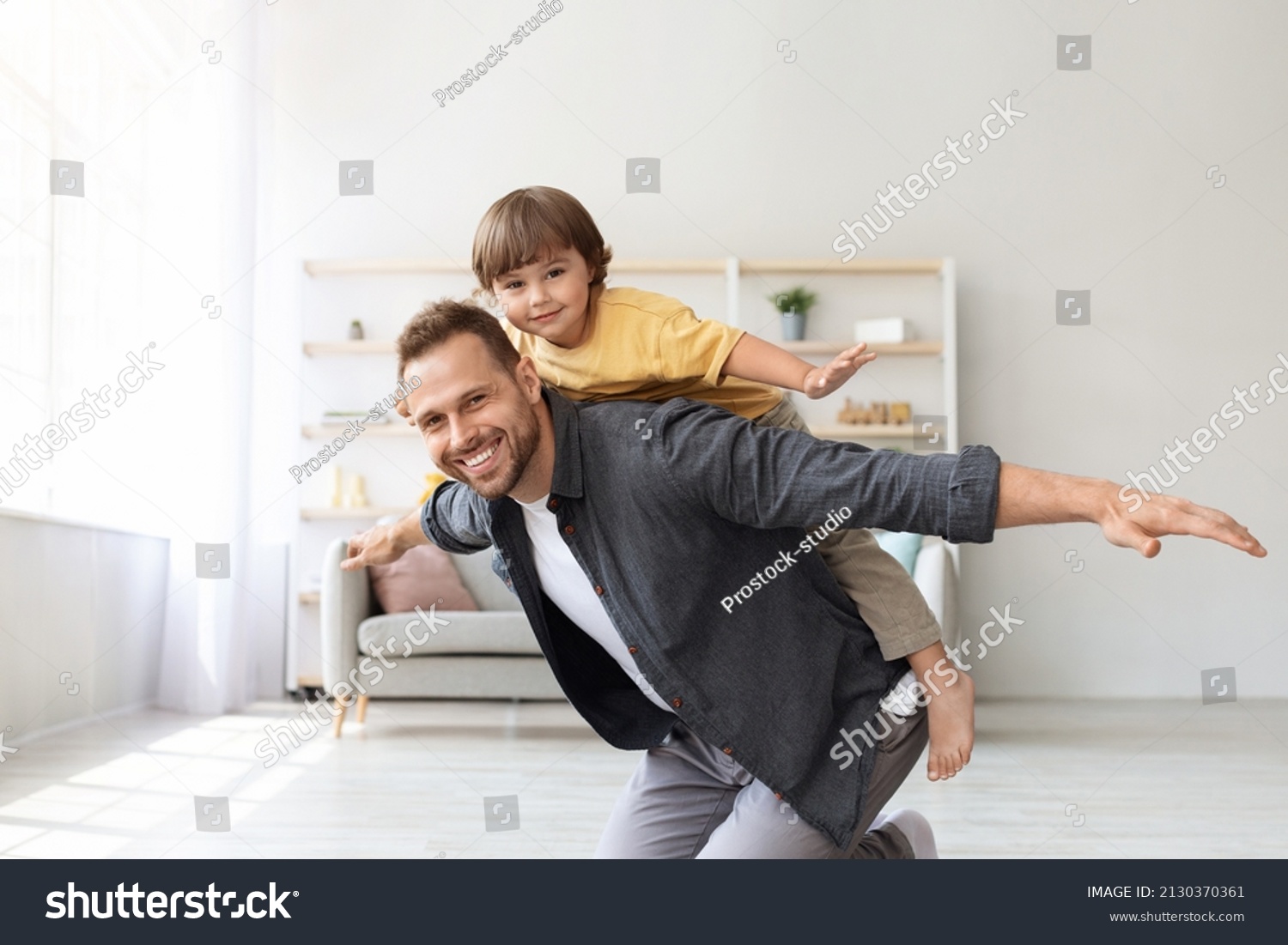 Games at home. Happy little boy riding daddy's back at home, kid playing plane together with father, smiling to camera at living room, free space #2130370361