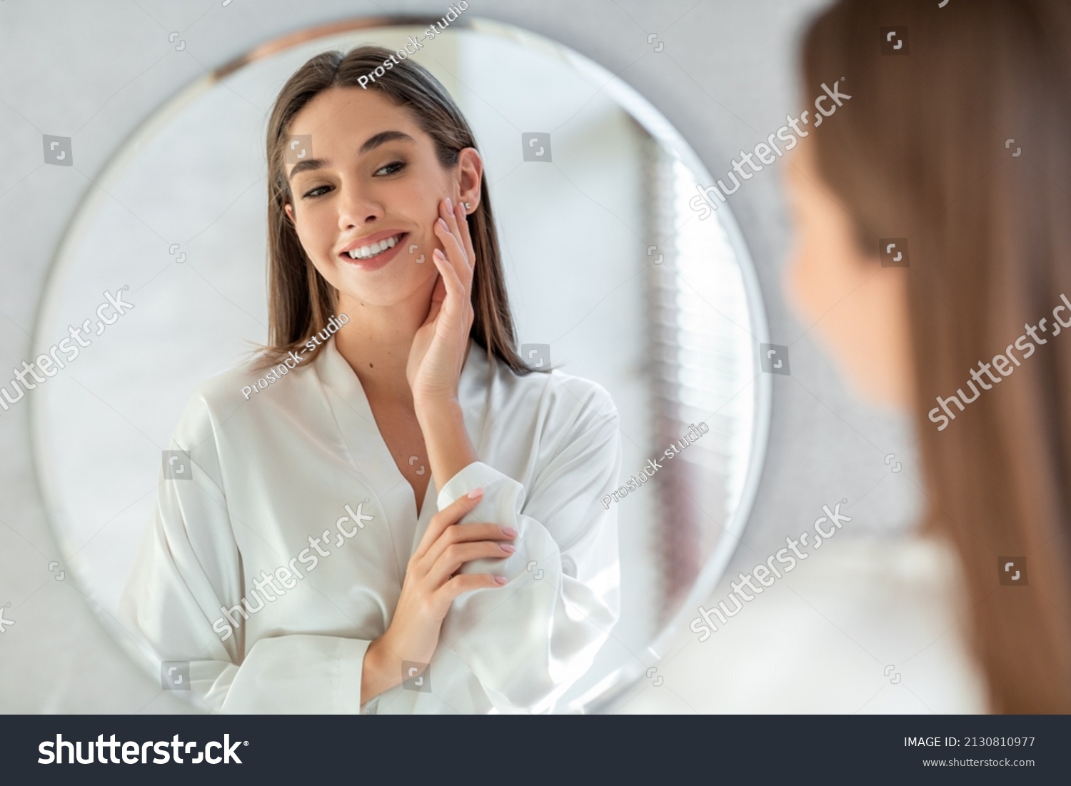 Self-Care Concept. Portrait Of Attractive Young Female Looking At Mirror, Beautiful Woman Wearing White Silk Robe Touching Soft Skin On Face And Smiling, Enjoying Her Reflection, Selective Focus #2130810977