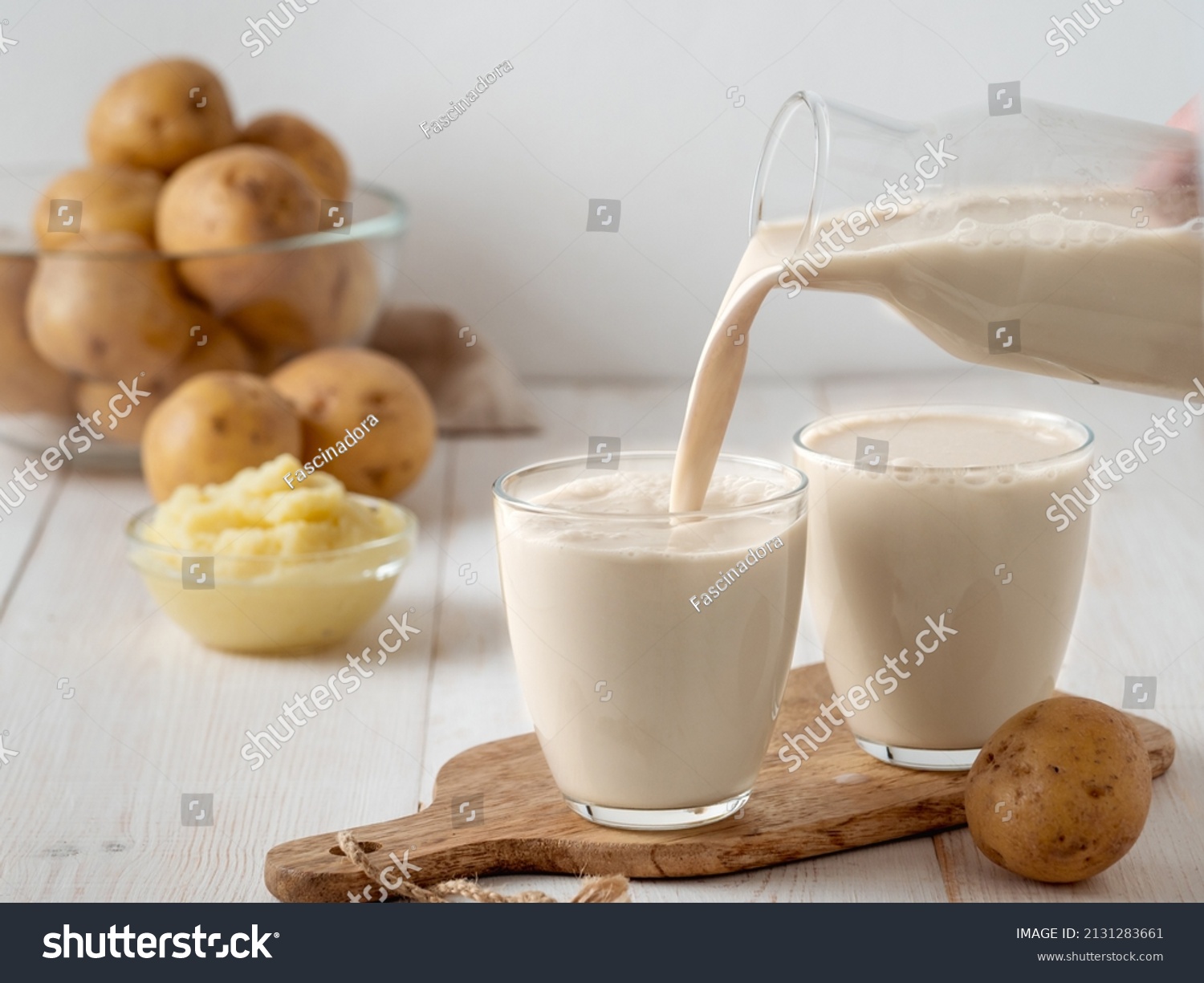 Potato milk pouring into glass on white wooden background. Pouring vegan milk in glass, with potato puree and potato tubers on background. Copy space. Home made potato milk made from boiled potatoes #2131283661