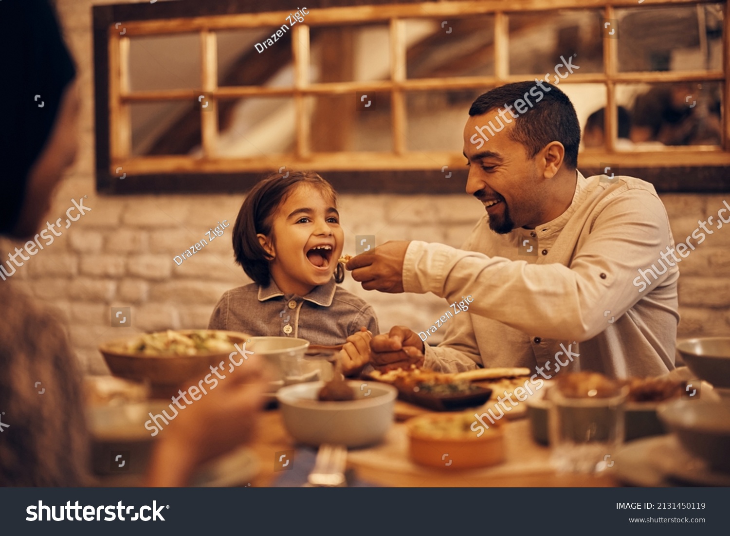 Happy Muslim little girl having fun while father is feeding her while having dinner at home on Ramadan.  #2131450119