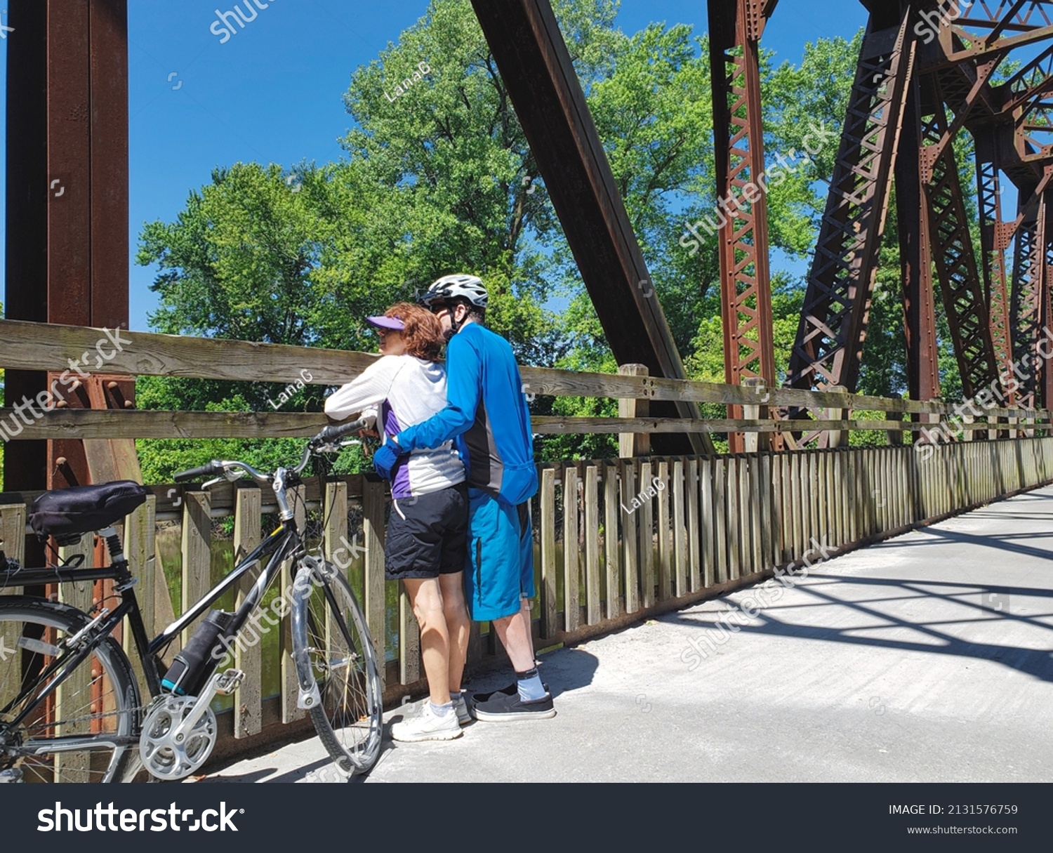 Older  bicyclists standing on old Midwestern bridge on clear summer day looking out; bicycle next the them #2131576759