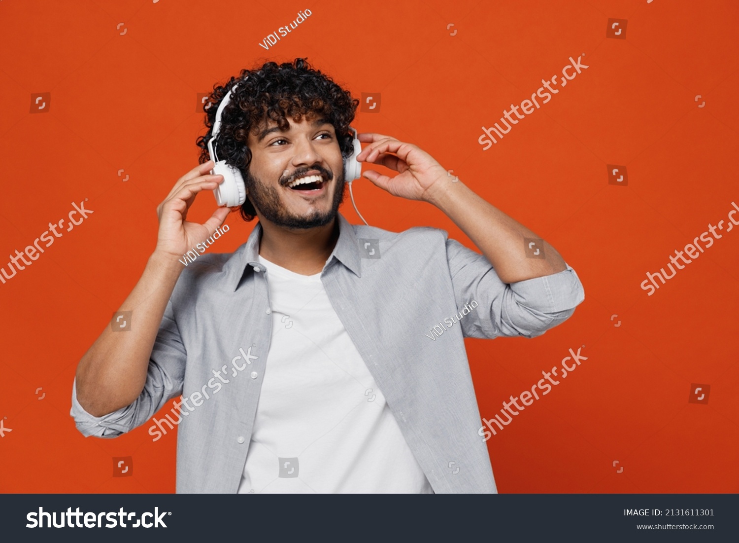 Cheerful exultant happy young bearded Indian man 20s years old wears blue shirt listen music in headphones dance have fun gesticulating hands relax isolated on plain orange background studio portrait #2131611301