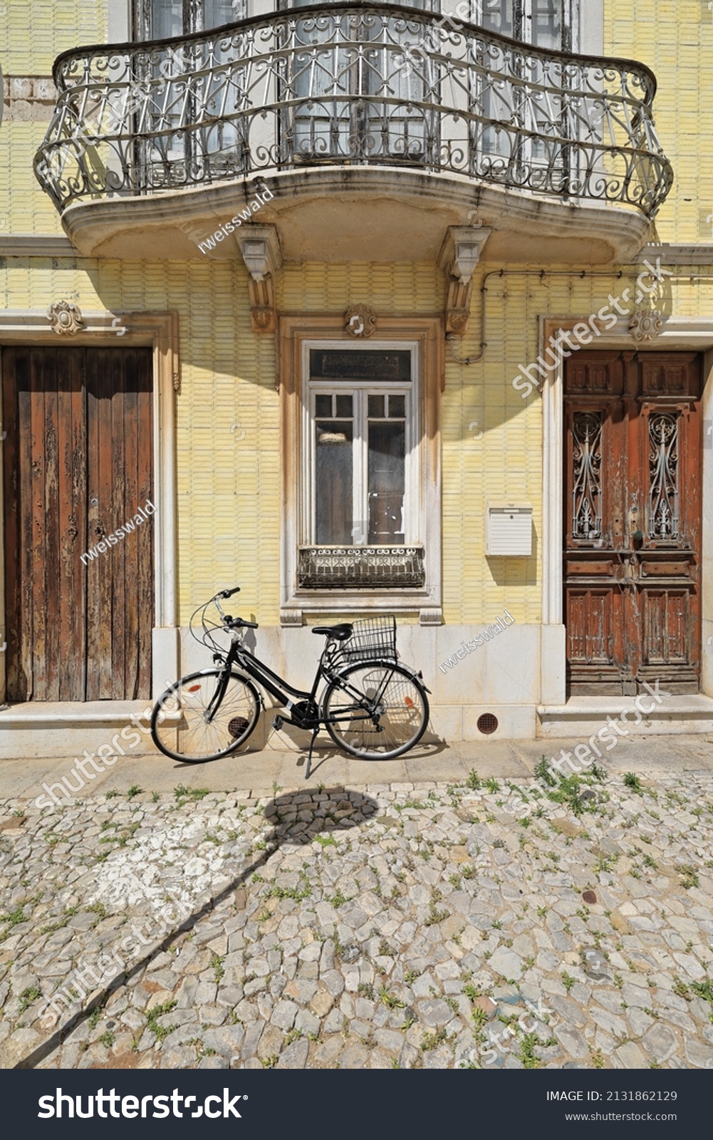 Yellow tile facade of old Neoclassical townhouse with black bicycle stopped at the door on the cobblestone sidewalk and chipped wooden door under metal railing balcony. Tavira-Algarve region-Portugal. #2131862129