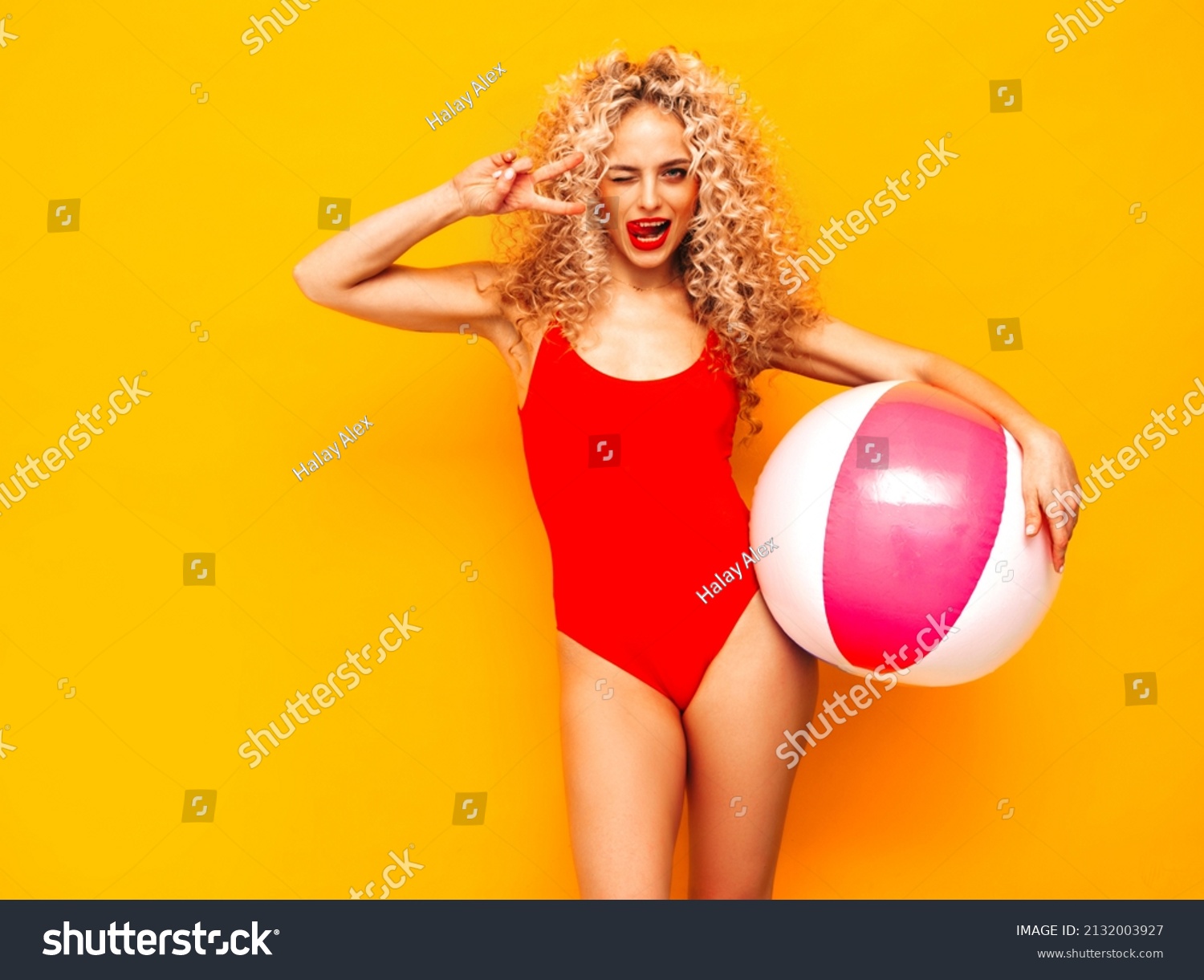 Young beautiful smiling woman posing near yellow wall in studio.Sexy model in red swimwear bathing suit.Positive female with curls hairstyle. Holding penny inflatable ball.Happy and cheerful #2132003927