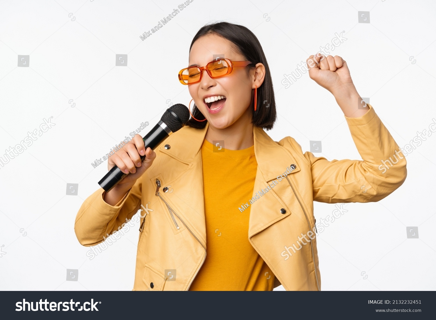 Stylish asian girl in sunglasses, singing songs with microphone, holding mic and dancing at karaoke, posing against white background #2132232451