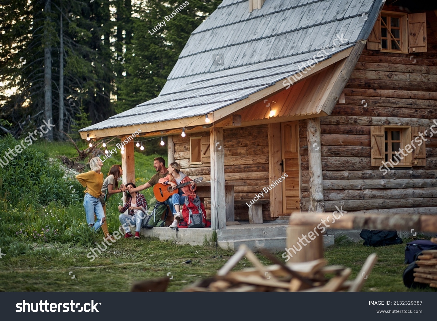 Young cheerful friends in front of wooden cottage on the terrace.  Girl playing guitar. Summertime garden celebration and fun. Friends, togetherness, fun, celebration concept. #2132329387