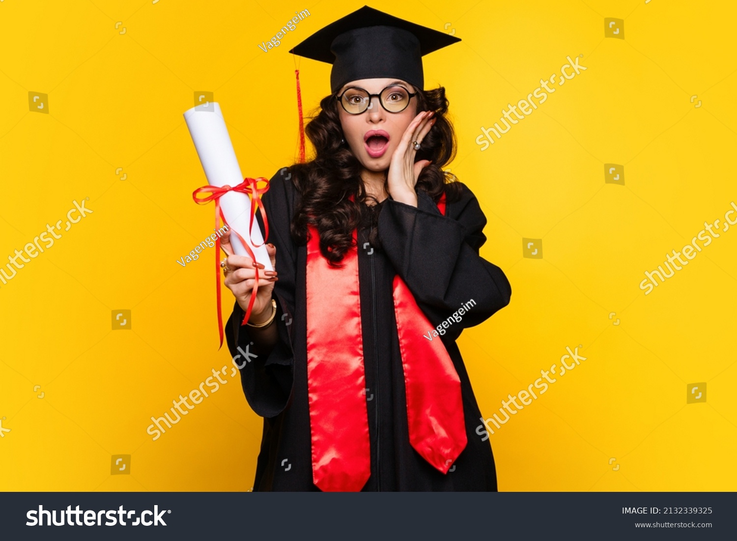Shocked Girl graduate in graduation hat and eyewear with diploma on yellow backdrop. pleasantly surprised young woman wearing graduation cap and ceremony robe holding Certificate. Education Concept #2132339325