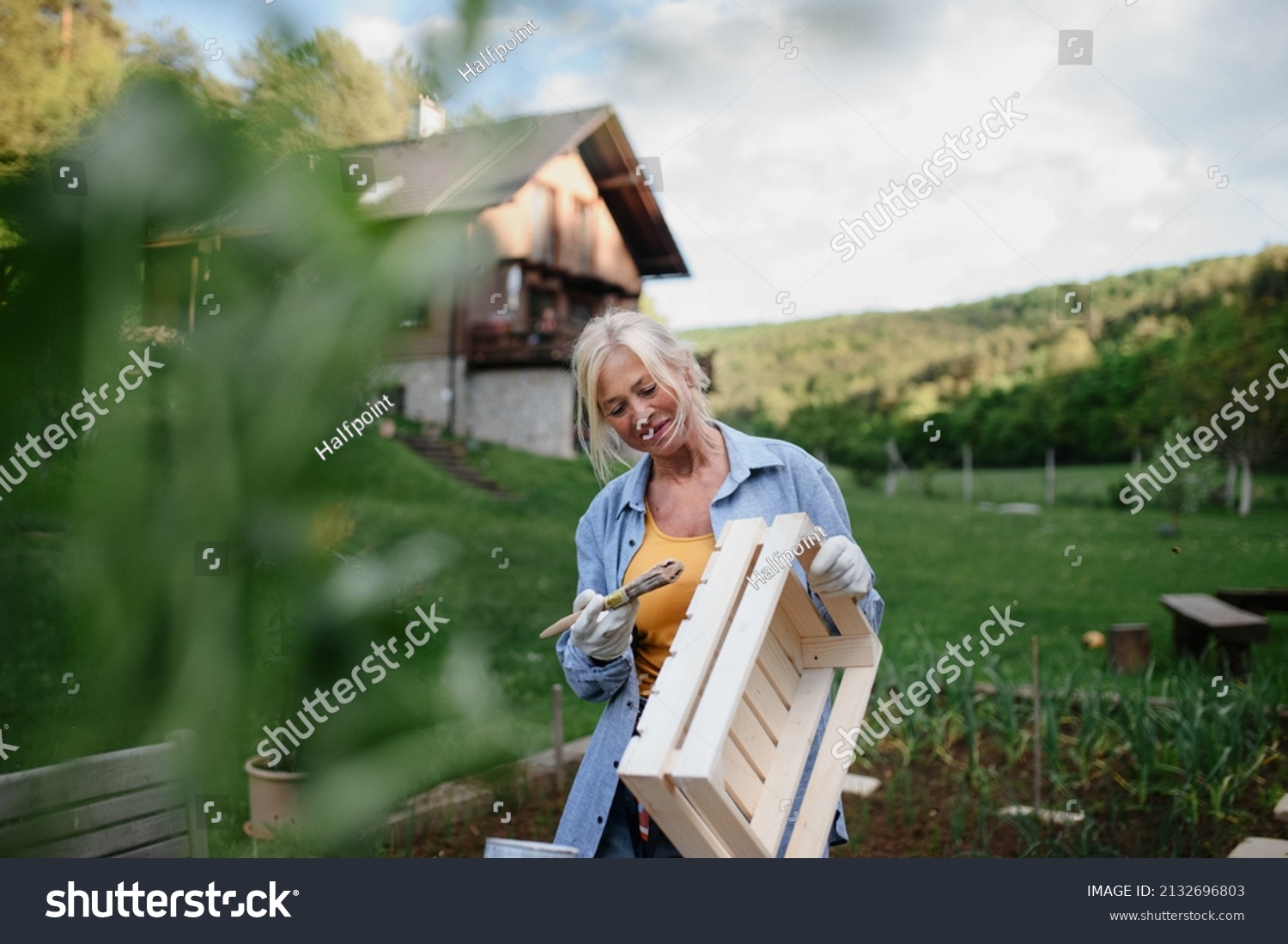 Senior woman with paintbrush impregnating wooden crate outdoors in garden. #2132696803