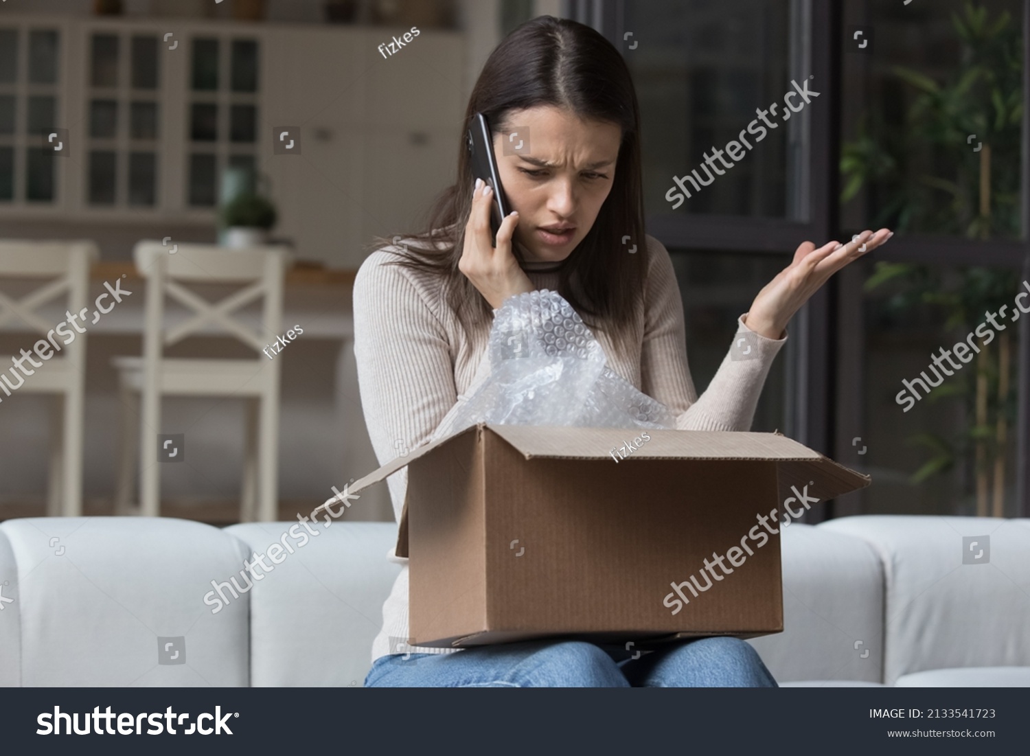 Dissatisfied angry woman, cheated client sit on sofa check received box, damaged or broken goods in parcel, talks to customers support, express complaints, looks annoyed. Bad delivery services concept #2133541723