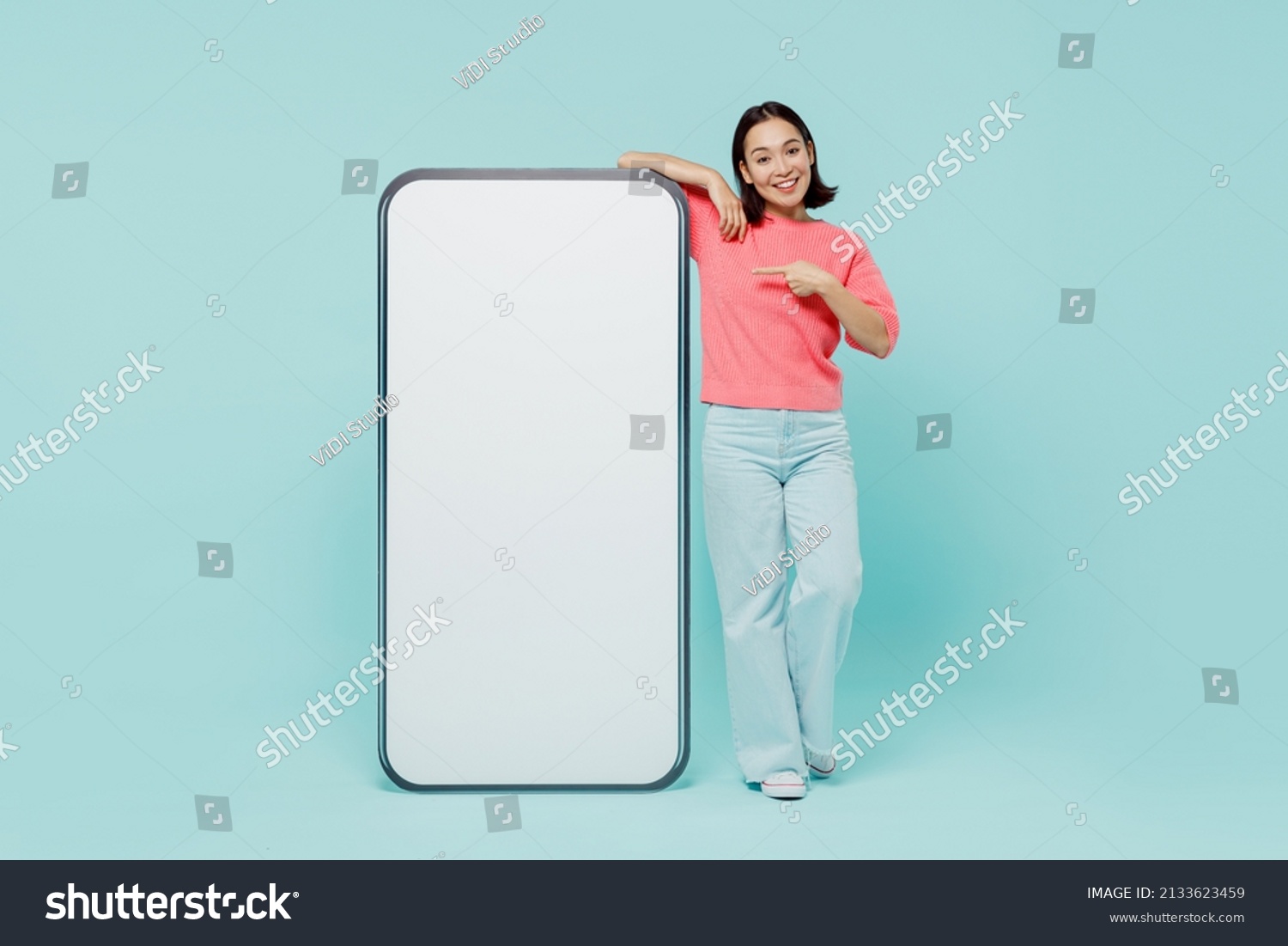 Full body young smiling happy woman of Asian ethnicity 20s in pink sweater point finger on big mobile cell phone with blank screen workspace area isolated on pastel plain light blue background studio #2133623459