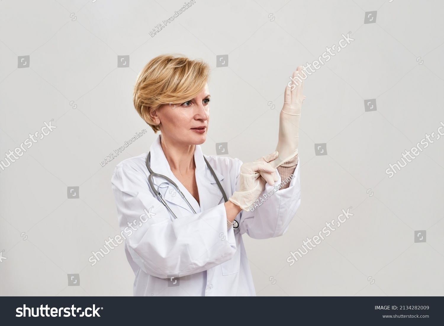 Adult female doctor putting latex glove on her hand. Caucasian woman with stethoscope wearing white coat. Concept of medical and health care. Isolated on white background. Studio shoot. Copy space #2134282009