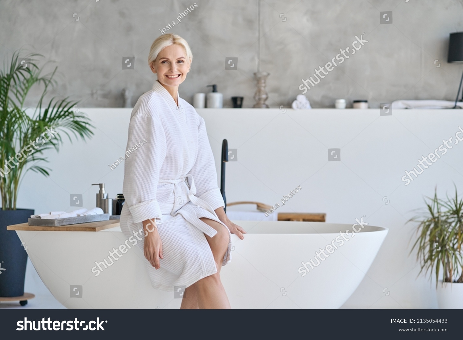 Wideshot of happy smiling beautiful middle aged woman in bathrobe at luxury spa hotel sitting on bath tube. Advertising of bodycare spa procedures antiage recreation skin care salon concept. #2135054433