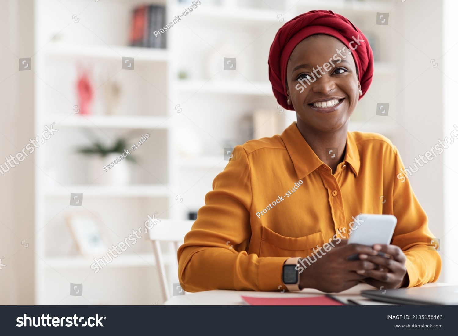 Positive beautiful young muslim black woman in red headscarf using brand new mobile phone, looking at copy space and smiling, using newest mobile application for business, home interior #2135156463