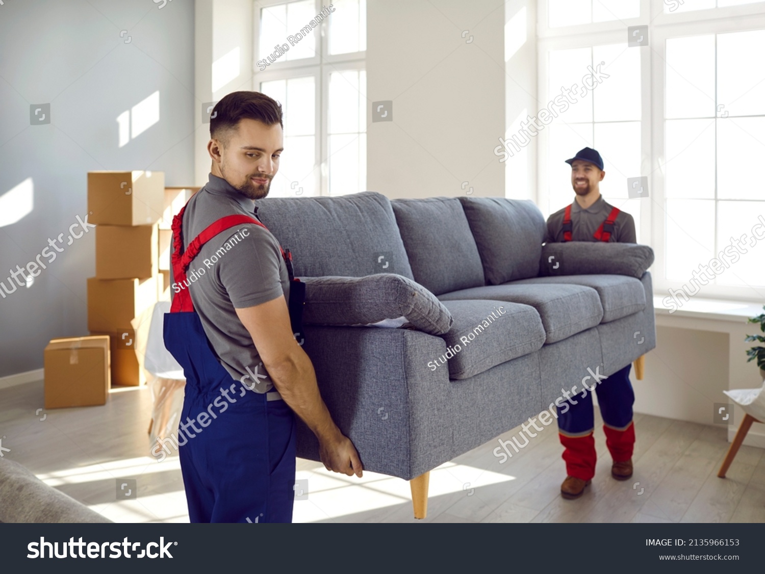 Two professional relocation service workers in overalls move sofa in customer's apartment. Movers carry sofa, cardboard boxes and assembling furniture. Moving and delivery company services. #2135966153