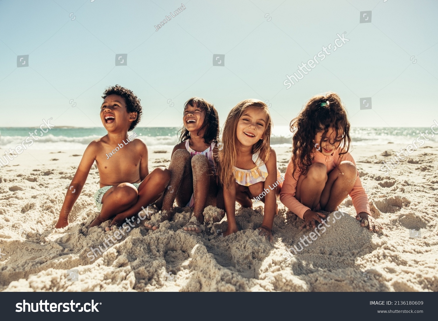 Four young friends laughing cheerfully while playing with sea sand at the beach. Group of adorable little kids having a good time together during summer vacation. #2136180609