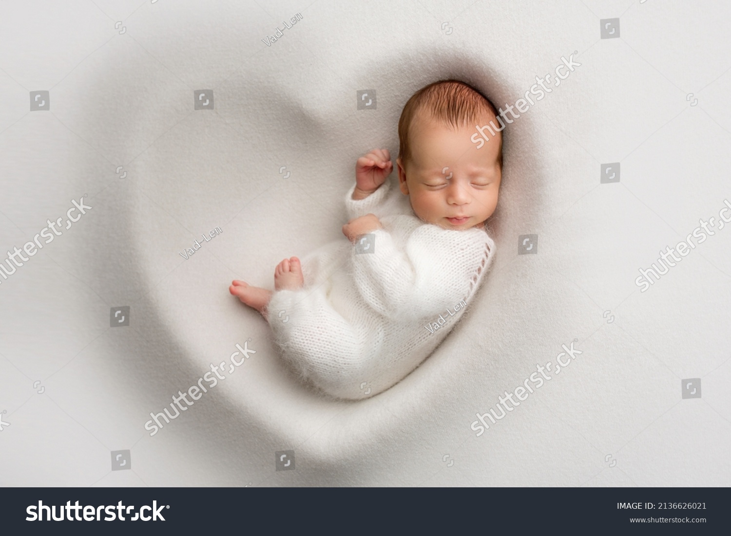 A sleeping newborn boy in the first days of life in a white soft cocoon with a knitted woolen white hat on a white background in the shape of a heart. Studio macro photography, portrait of a newborn. #2136626021