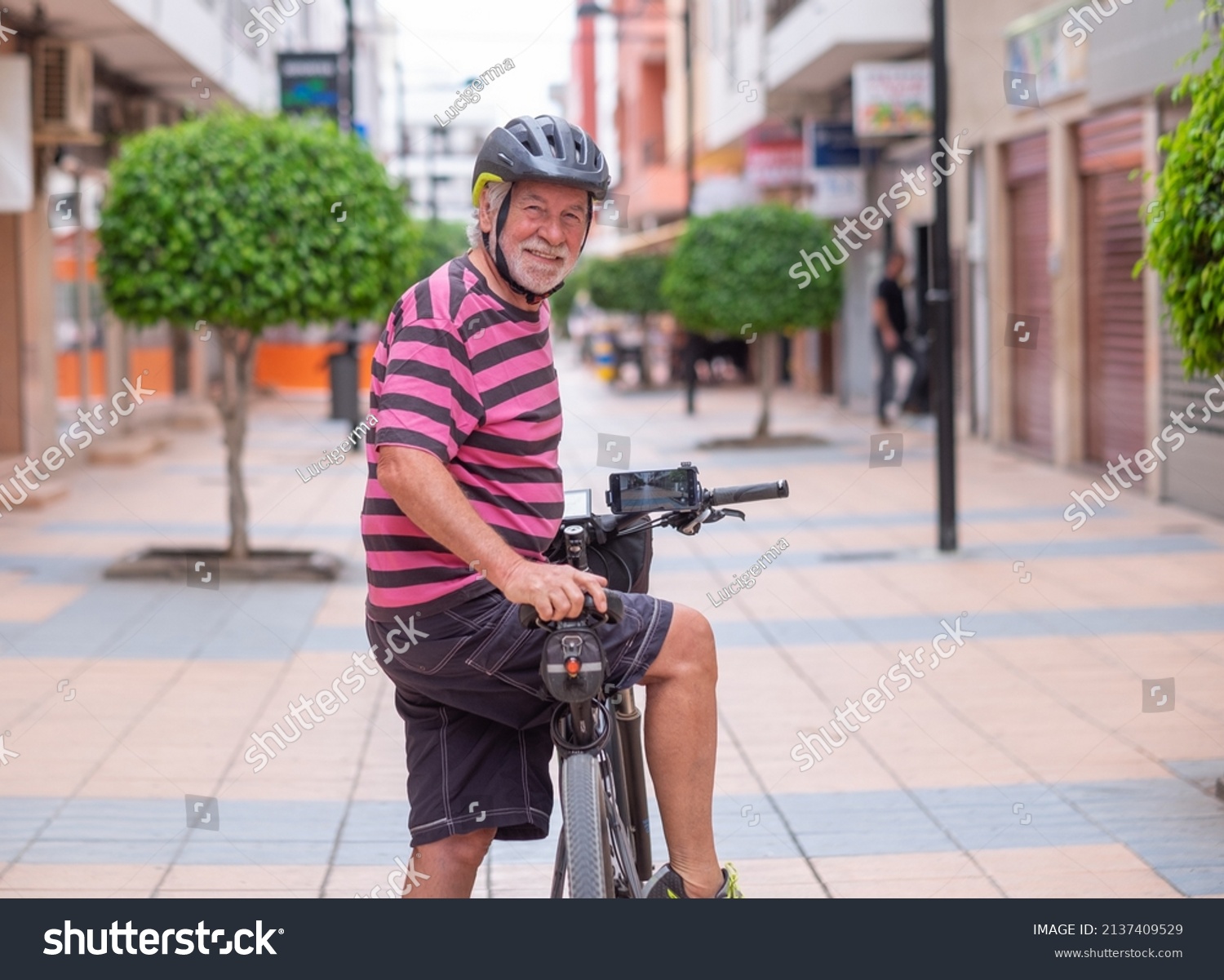 Senior smiling cyclist man wearing helmet cycling his electric bicycle in urban street stops looking back at camera. Active elderly grandfather enjoying a healthy lifestyle and freedom #2137409529