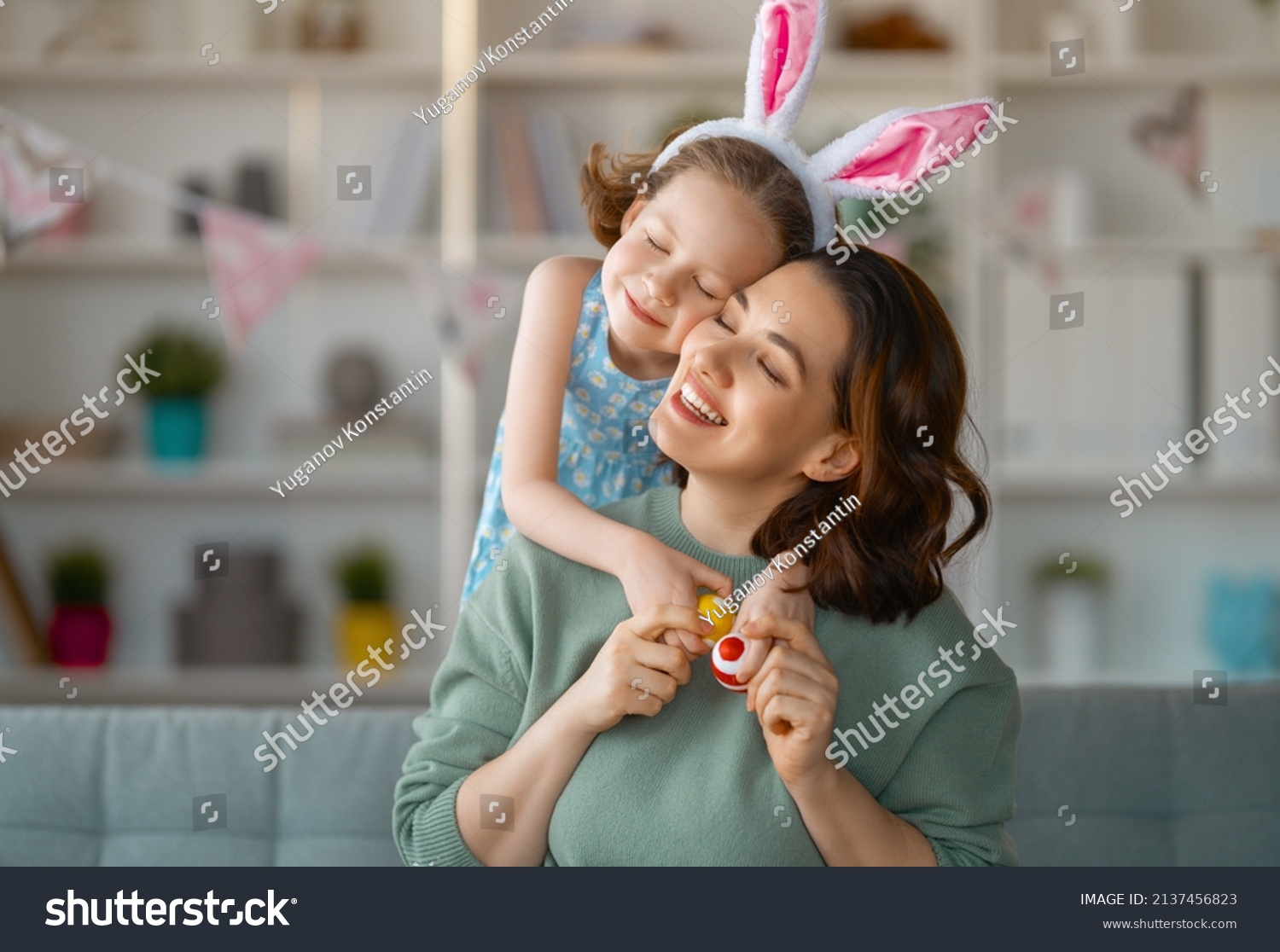 Happy holiday! Mother and her daughter with painting eggs. Family celebrating Easter. Cute little child girl is wearing bunny ears. #2137456823