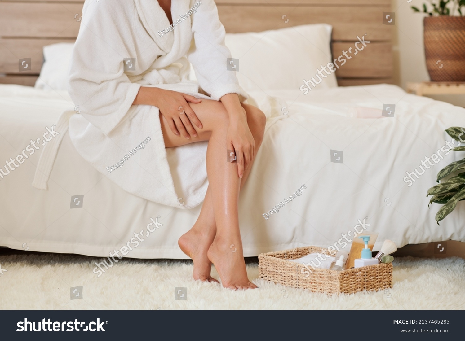 Young woman in bathrobe sitting on bed and applying body oil after shower #2137465285