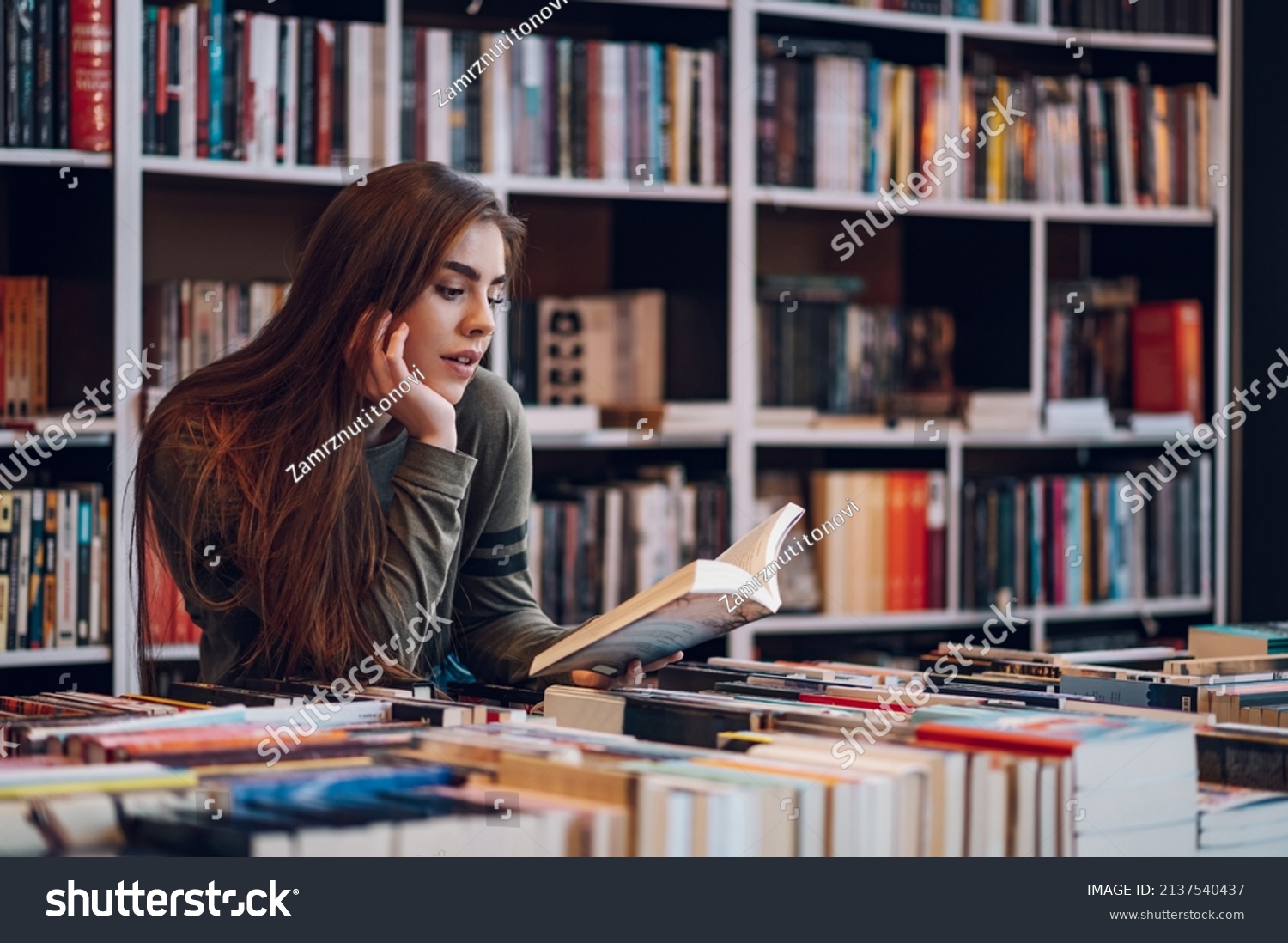 Beautiful young woman buying books at a bookstore and reading one. Geeky woman reading a book with a bookshelves in background. #2137540437