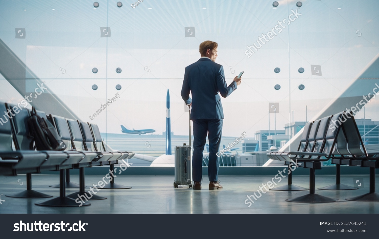 Airport Terminal: Businessman with Rolling Suitcase Walks, Uses Smartphone App for e-Business. Back View Silhouette of Traveling Man Waits for Flight in Boarding Lounge of Airline Hub with Airplanes #2137645241