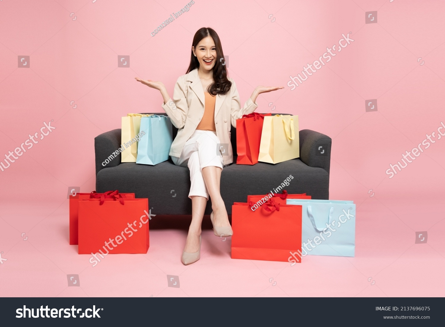 Full length portrait of Excited Asian woman sitting on sofa with shopping bags isolated on pink background, Shopper or shopaholic concept #2137696075
