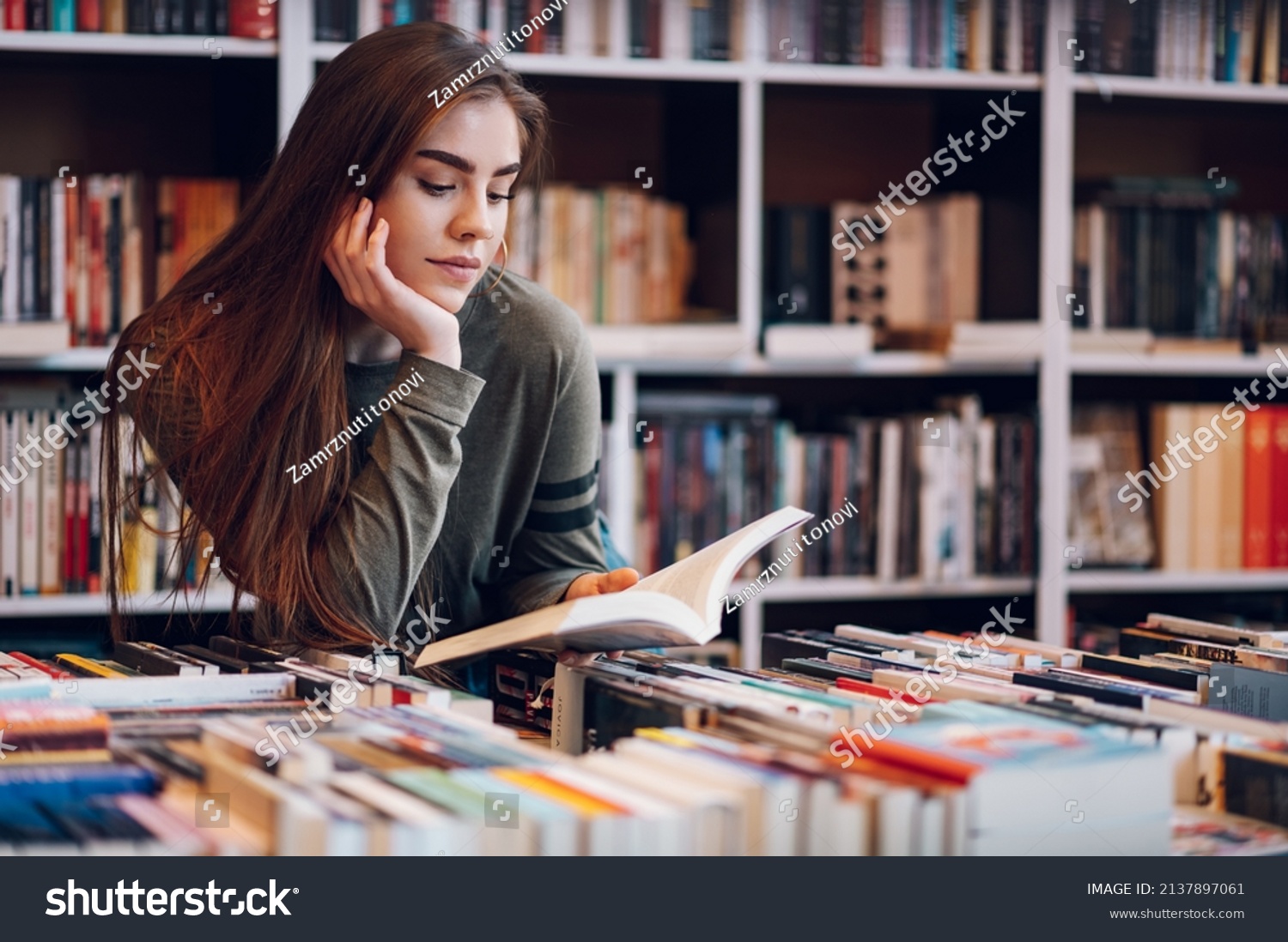 Young female customer reading a book in bookstore while buying some good literature. Woman picking a book to read. #2137897061
