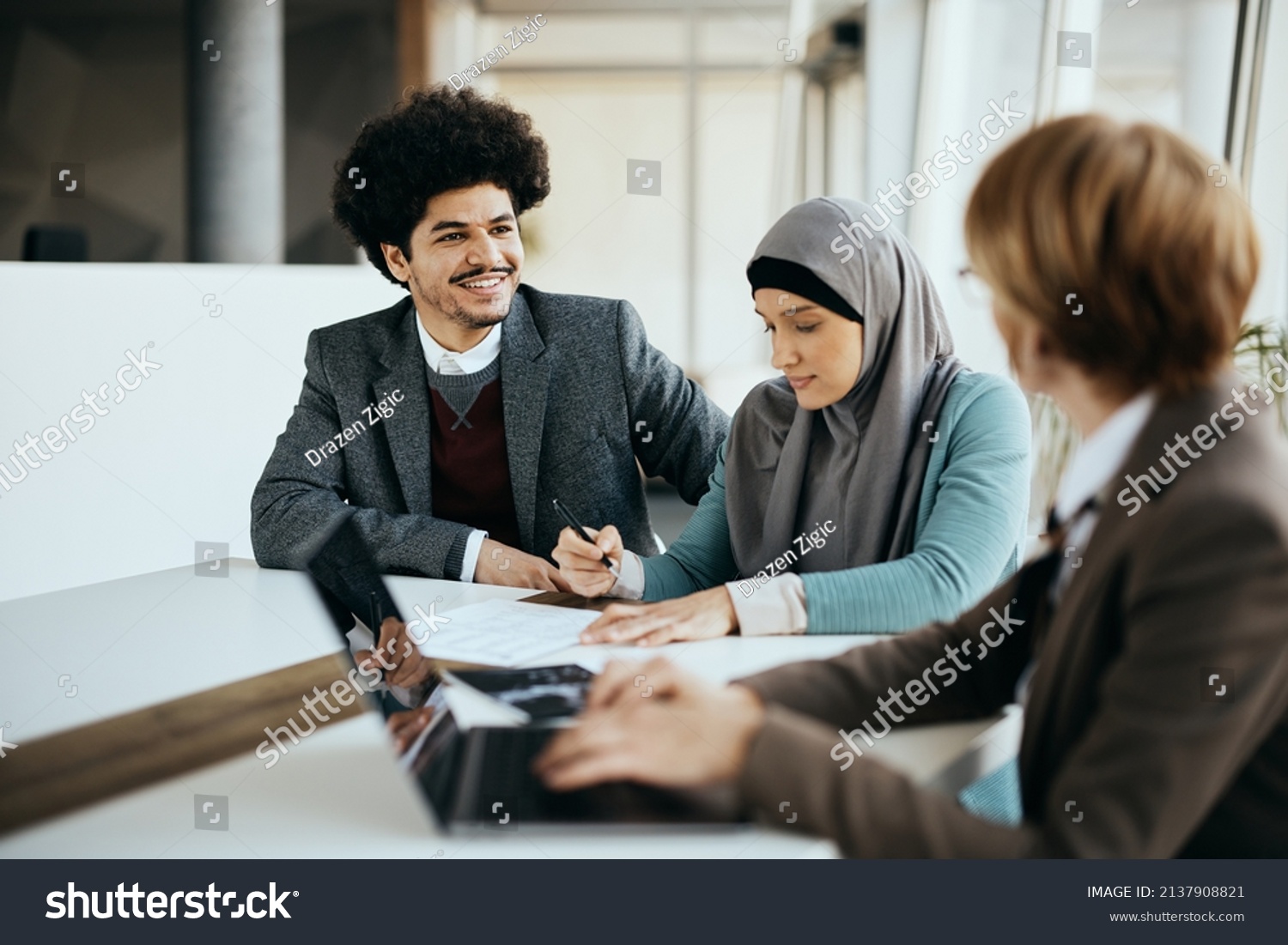 Happy Muslim man talking to financial advisor while his wife is signing paperwork during the meeting in the office.  #2137908821