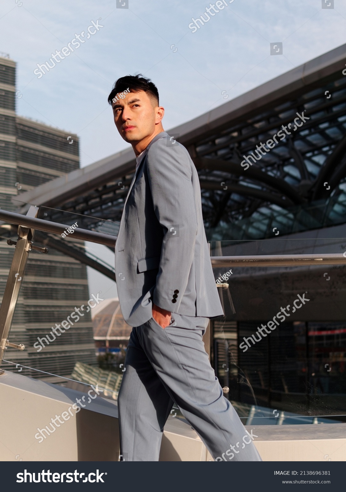 Portrait of handsome Chinese young man in light blue suit and white undershirt walking and looking away with modern city buildings background in sunny day, side view of confident businessman. #2138696381