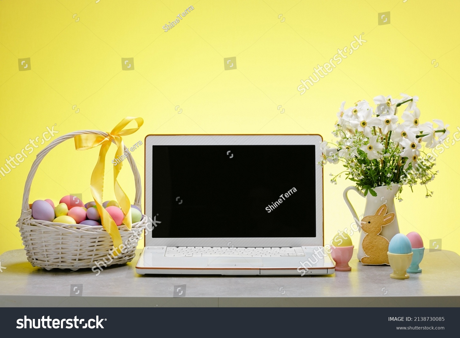 A laptop with a blank screen on a table festively decorated for the Easter holiday. Adorned working place with flowers and a basket with colorful eggs #2138730085
