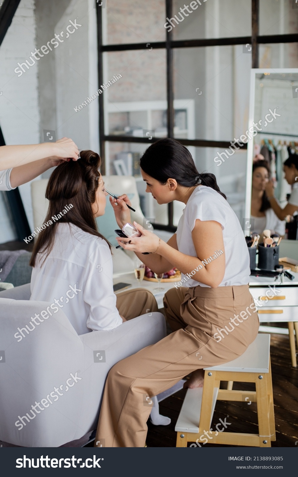 Woman makeup artist and hairdresser getting ready female to event wedding or important meeting. Group of feminine friends preparing cosmetic beauty care hair face procedure at home bedroom interior #2138893085