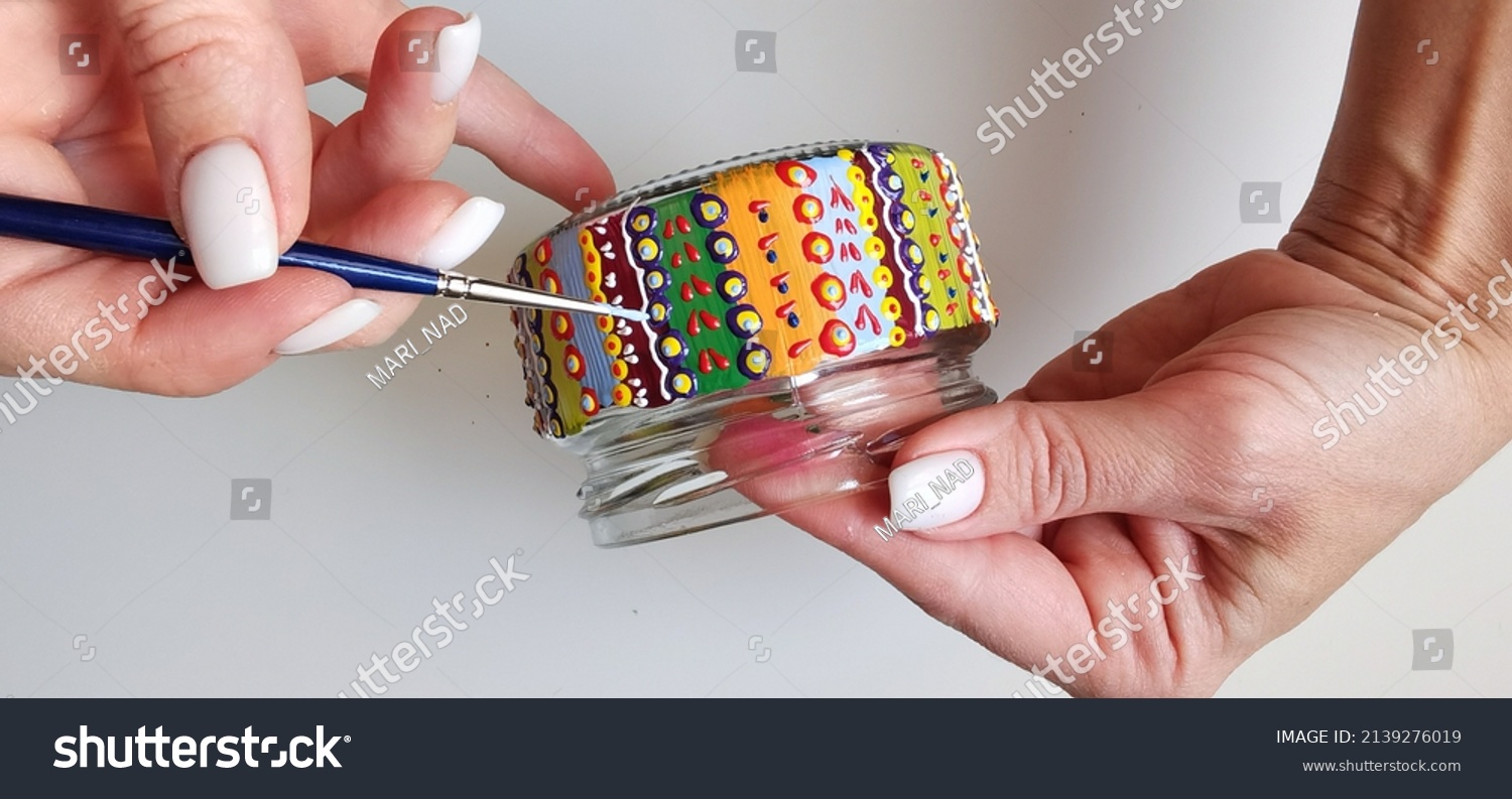 A woman holds a jar in her hands. Spot painting on glass, workshop. Drawing lesson for children in peacetime. Decorates the jar. Multicolored stained glass paints.Ideas for the decoration of objects.  #2139276019