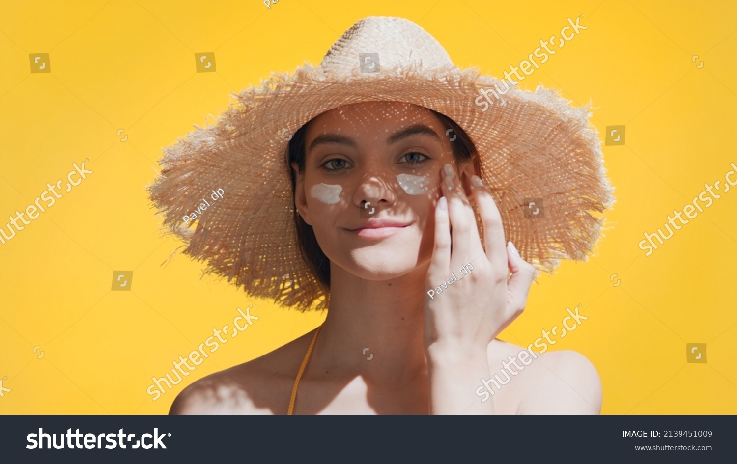 Young good-looking brunette Caucasian woman in a straw hat puts spf cream on her cheeks, enjoys the sun and smiles for the camera against yellow background | Spf cream concept #2139451009
