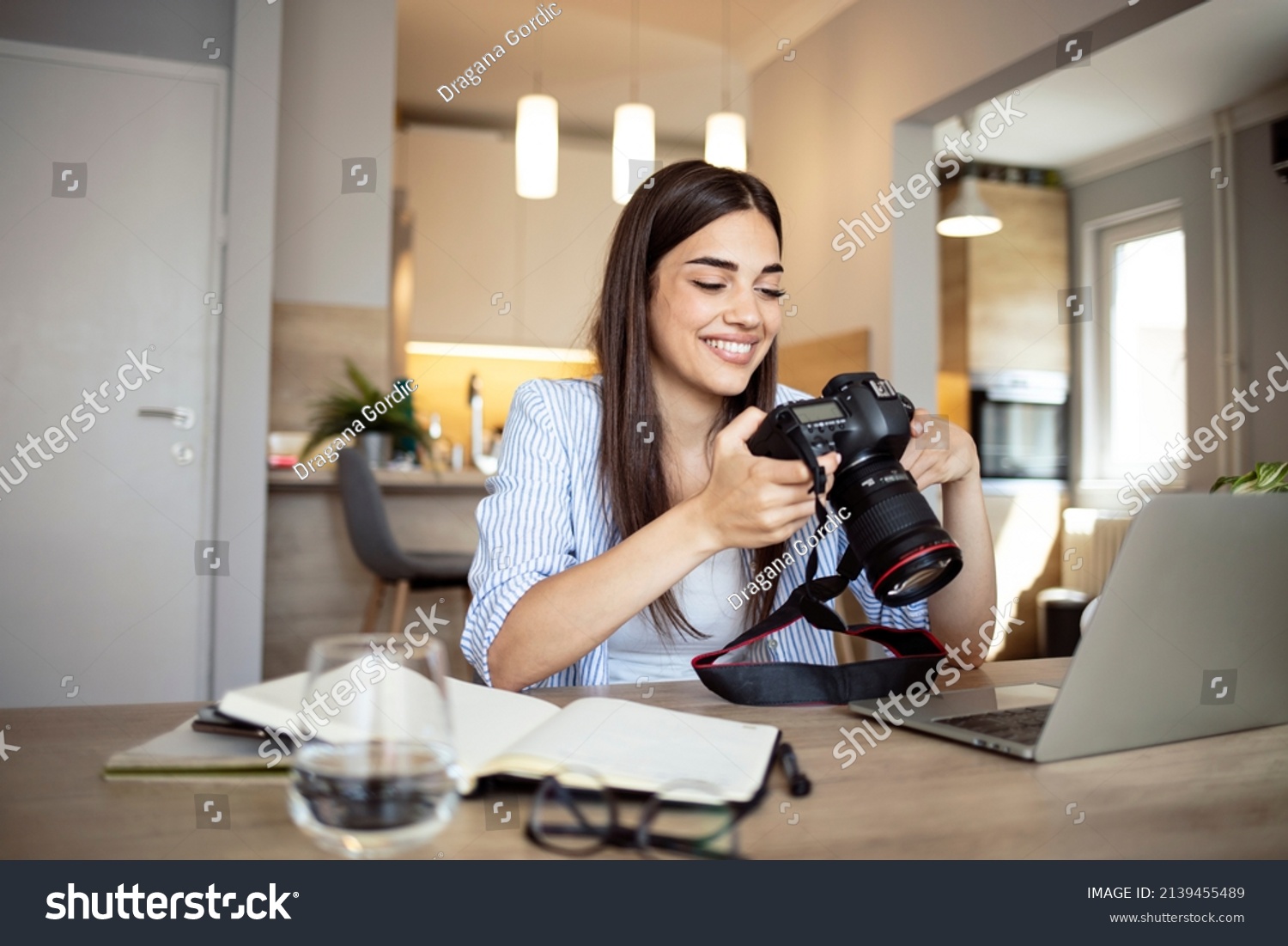 Happy young photographer holding a dslr camera in her home office. Female photographer smiling cheerfully while working at her desk. Creative female freelancer working on a new project. #2139455489