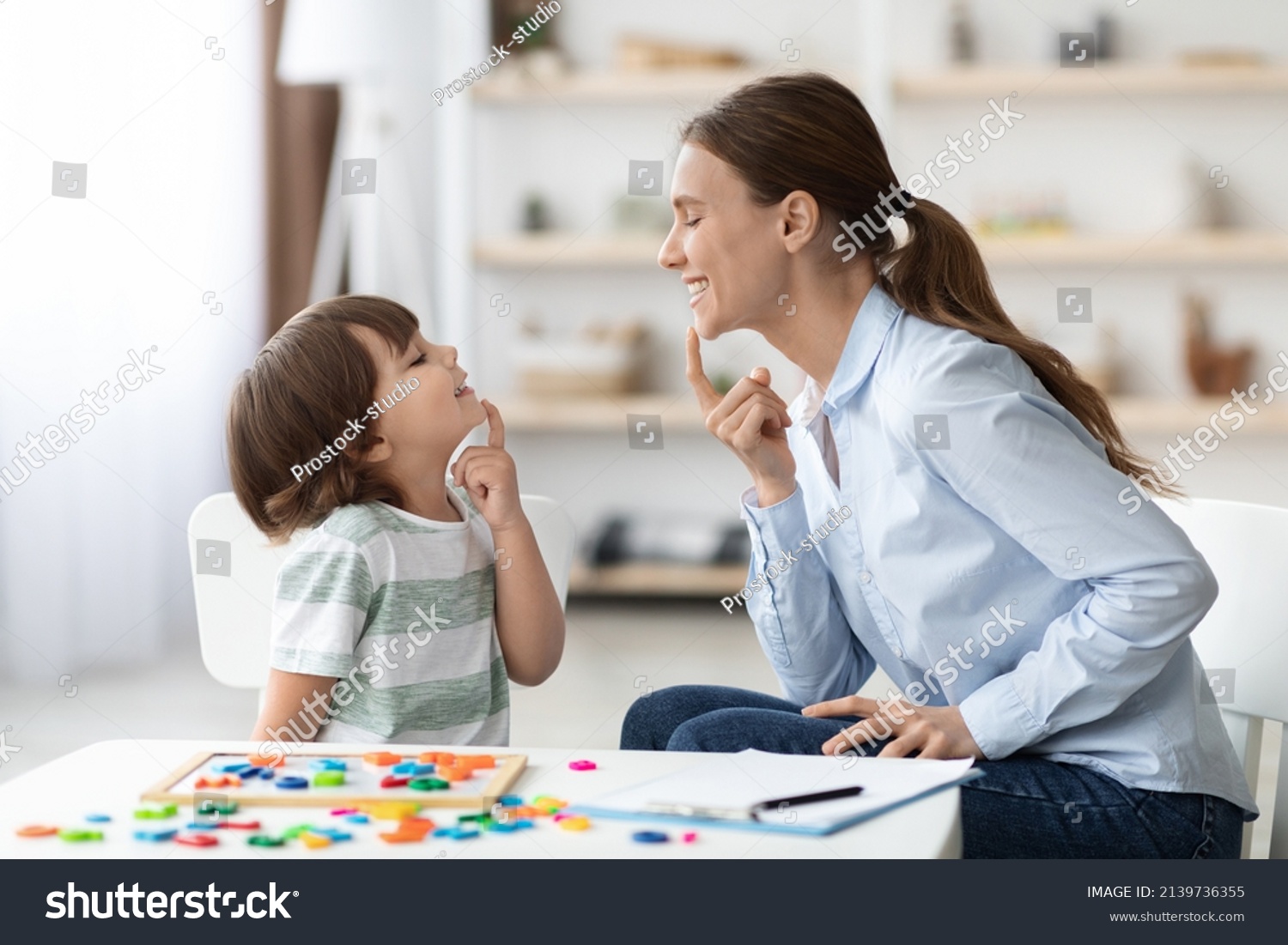Speech training for kids. Professional woman specialist training with little boy at cabinet, teaching him right articulation exercises, side view #2139736355