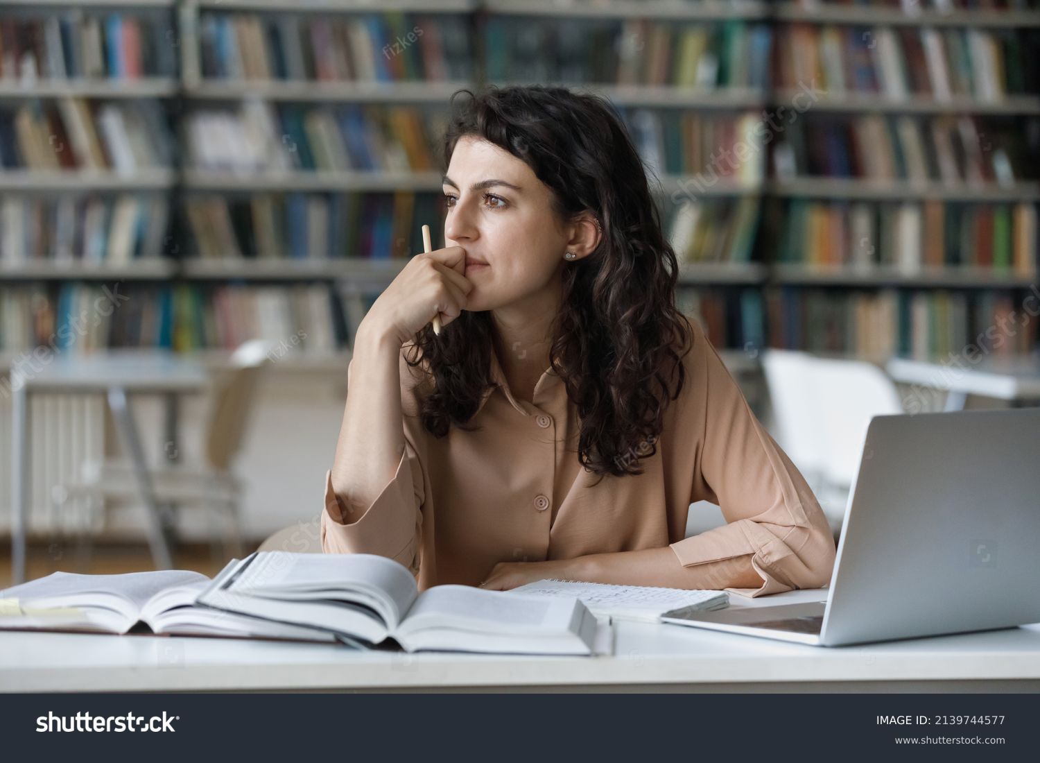 Young student girl sit at table with textbooks and laptop staring aside, studying alone in library, looks pensive and thoughtful search solution, prepare for exam, makes task feels confused or puzzled #2139744577