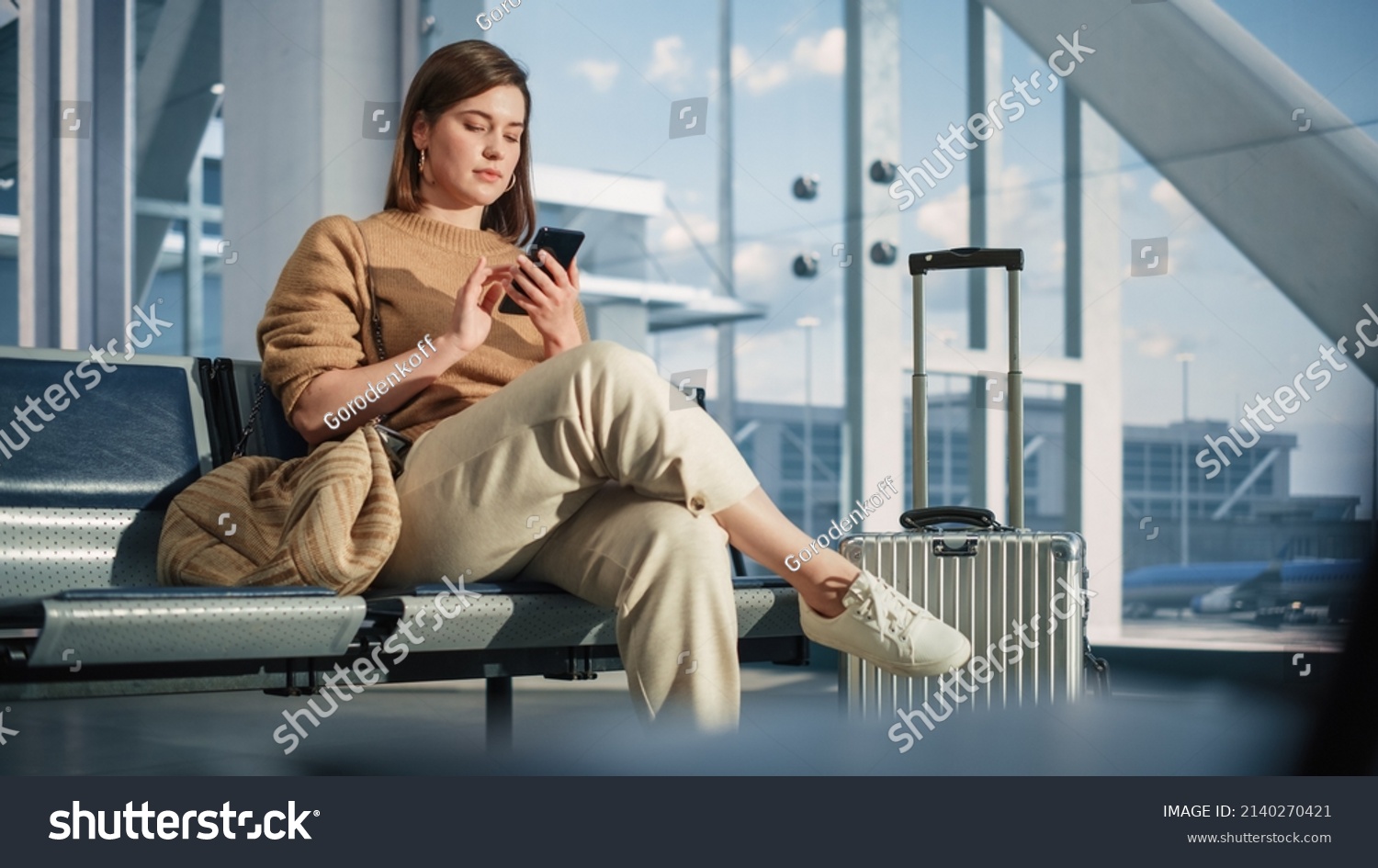 Airport Terminal: Woman Waits for Flight, Uses Smartphone, Browse Internet, Social Media, Online Shopping. Traveling Female Remote Work Online on Mobile Phone in a Boarding Lounge of Airline Hub #2140270421