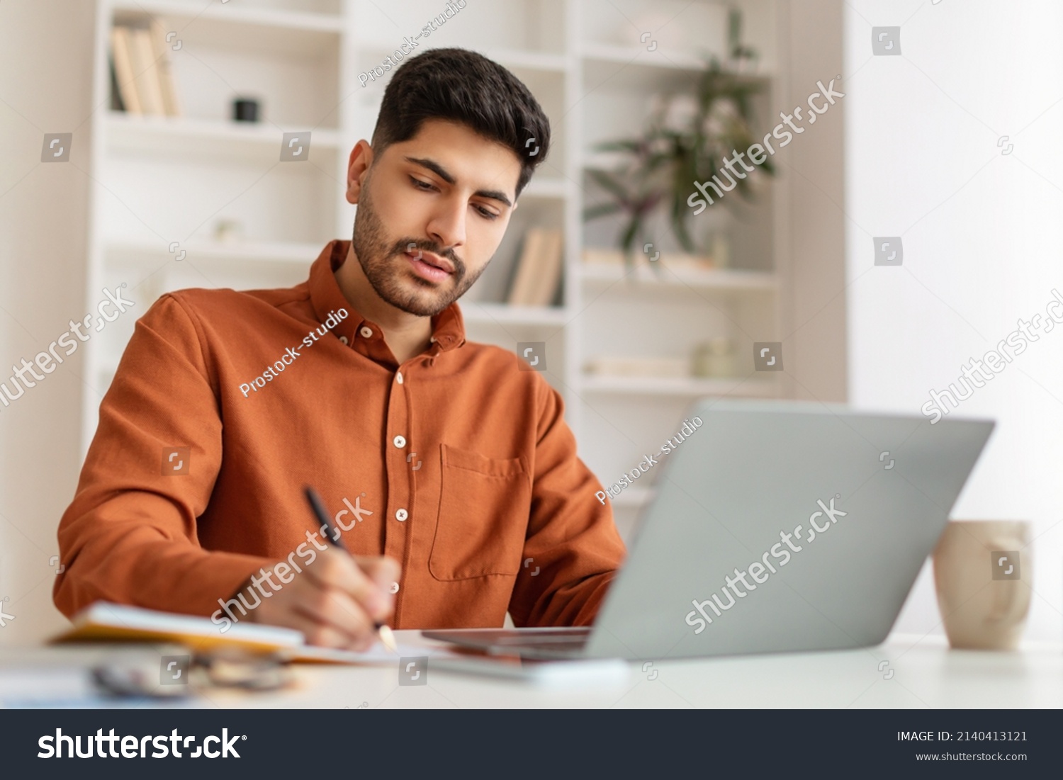 Focused young Middle Eastern man sitting at desk working on laptop taking notes in notebook, busy millennial male studying online, watching webinar using computer and writing check list #2140413121