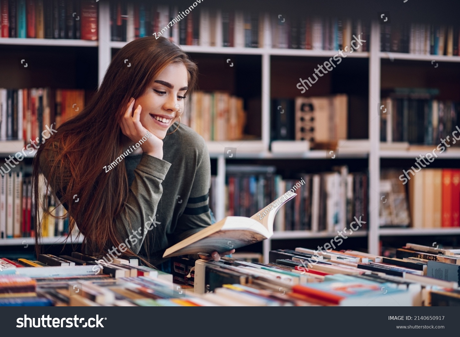 Portrait of a pretty smiling girl reading book while buying in a bookstore. Choosing a good book to buy in a bookstore. Woman interested in a book. #2140650917