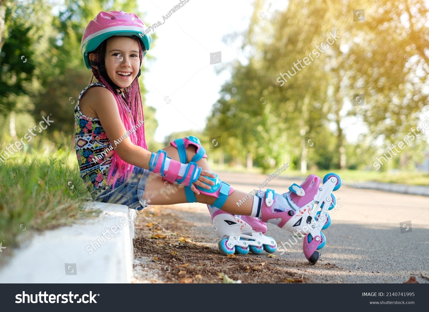 Laughing loudly, a funny girl with Afro-pigtails and wearing sports protective gloves and a helmet sat down to rest on the sidewalk after roller skating. A child's favorite hobby is roller skates. #2140741995