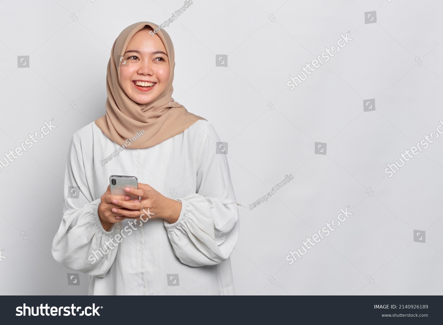 Smiling young Asian Muslim woman using a mobile phone and looking away isolated over white background #2140926189