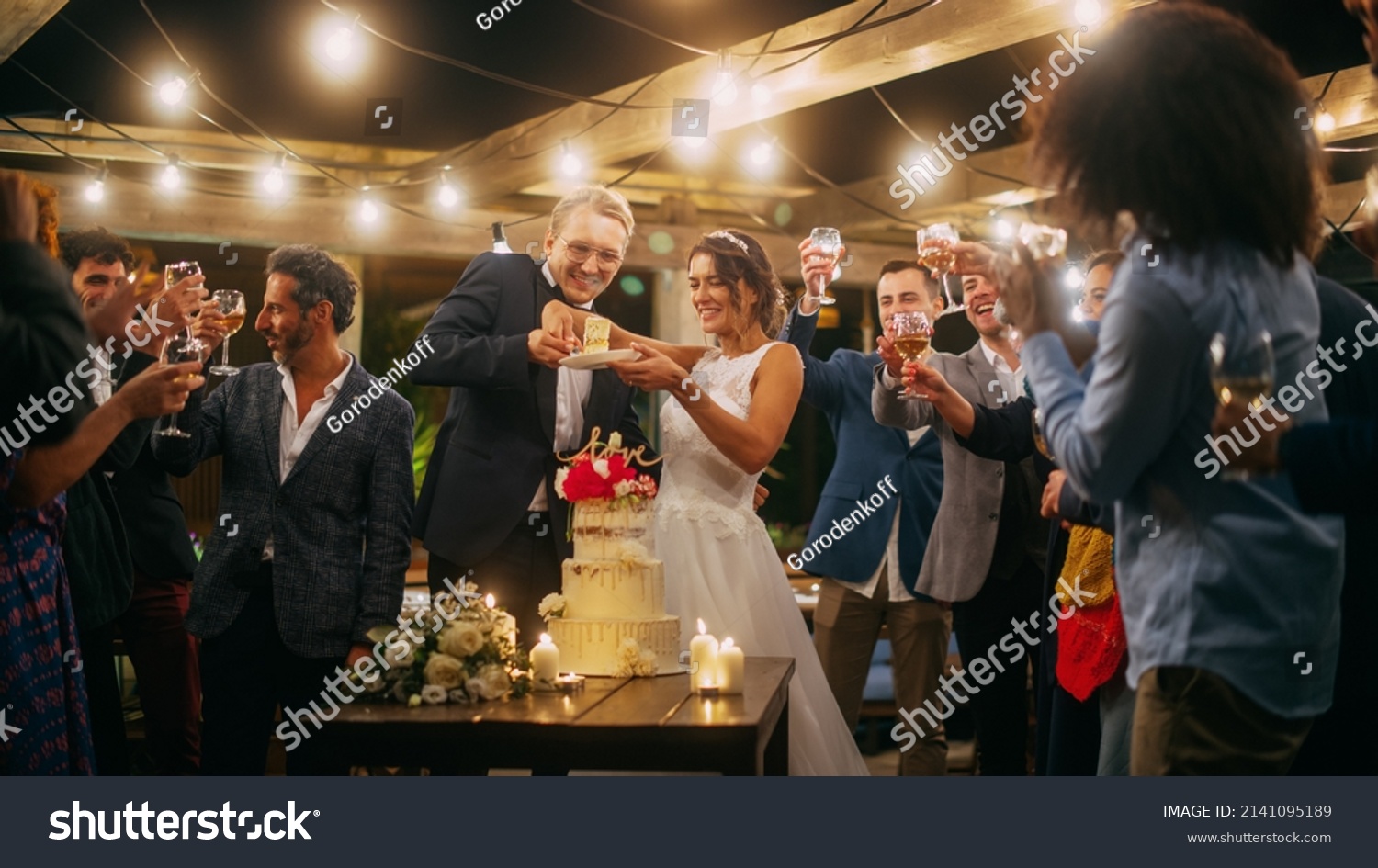 Beautiful Bride and Groom Celebrate Wedding at an Evening Reception Party with Multiethnic Friends. Married Couple Standing at a Dinner Table, Kiss and Cut Wedding Cake. #2141095189