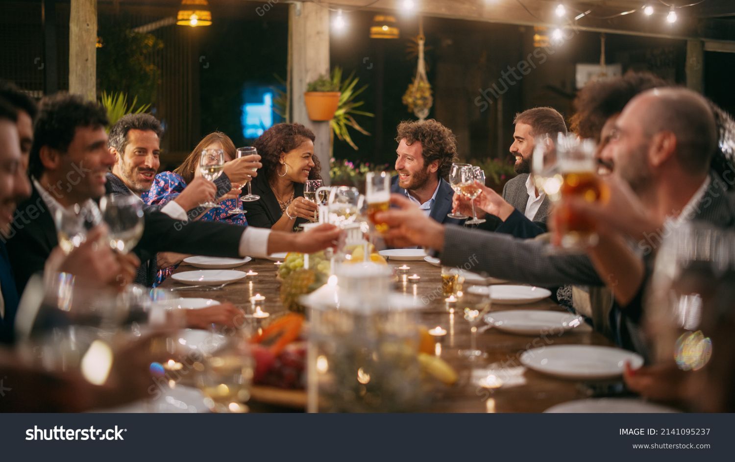 Big Dinner Party with a Small Crowd of Multiethnic Diverse Friends Celebrating at a Restaurant. Beautiful Happy Hosts Propose a Toast and Raise Wine Glasses while Sitting at a Table in the Evening. #2141095237