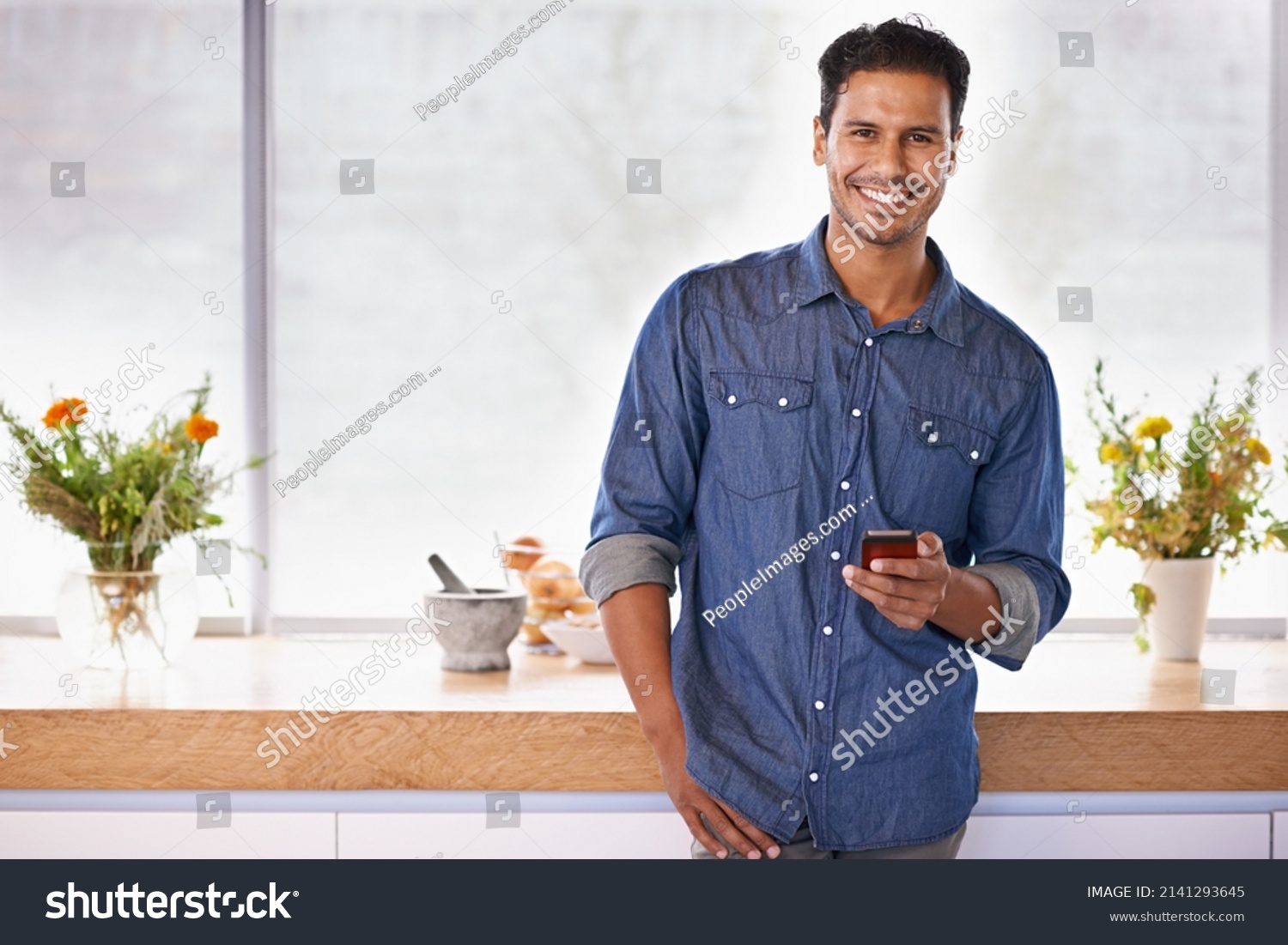Maybe I should invite some friends over for dinner. Portrait of a handsome holding a mobile phone while standing in the kitchen. #2141293645