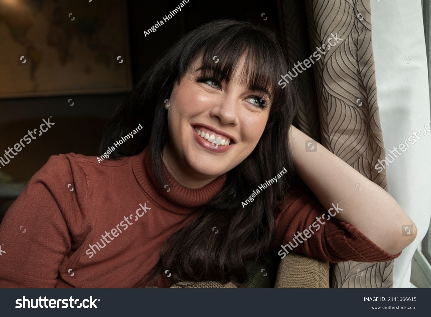 Portrait of a caucasian woman in her 30s smiling while sitting on the couch near a window.  #2141666615
