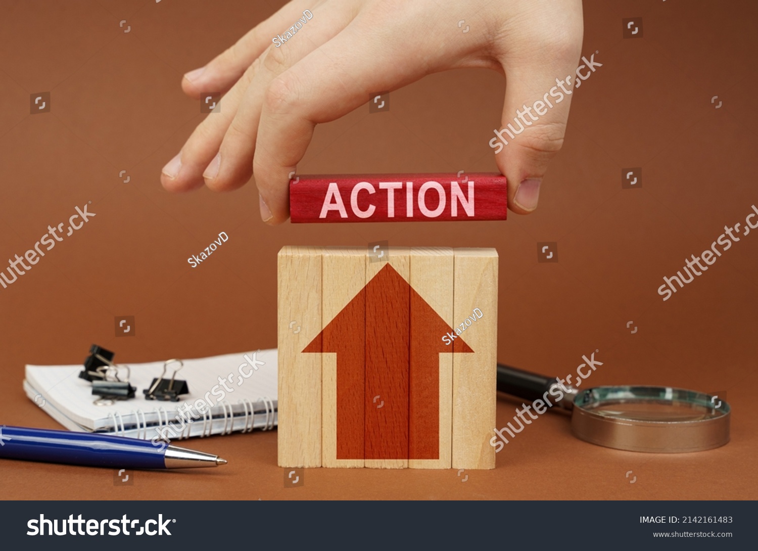 Business concept. On a brown surface are office items, wooden blocks, in the hand is a red block with the inscription - ACTION #2142161483