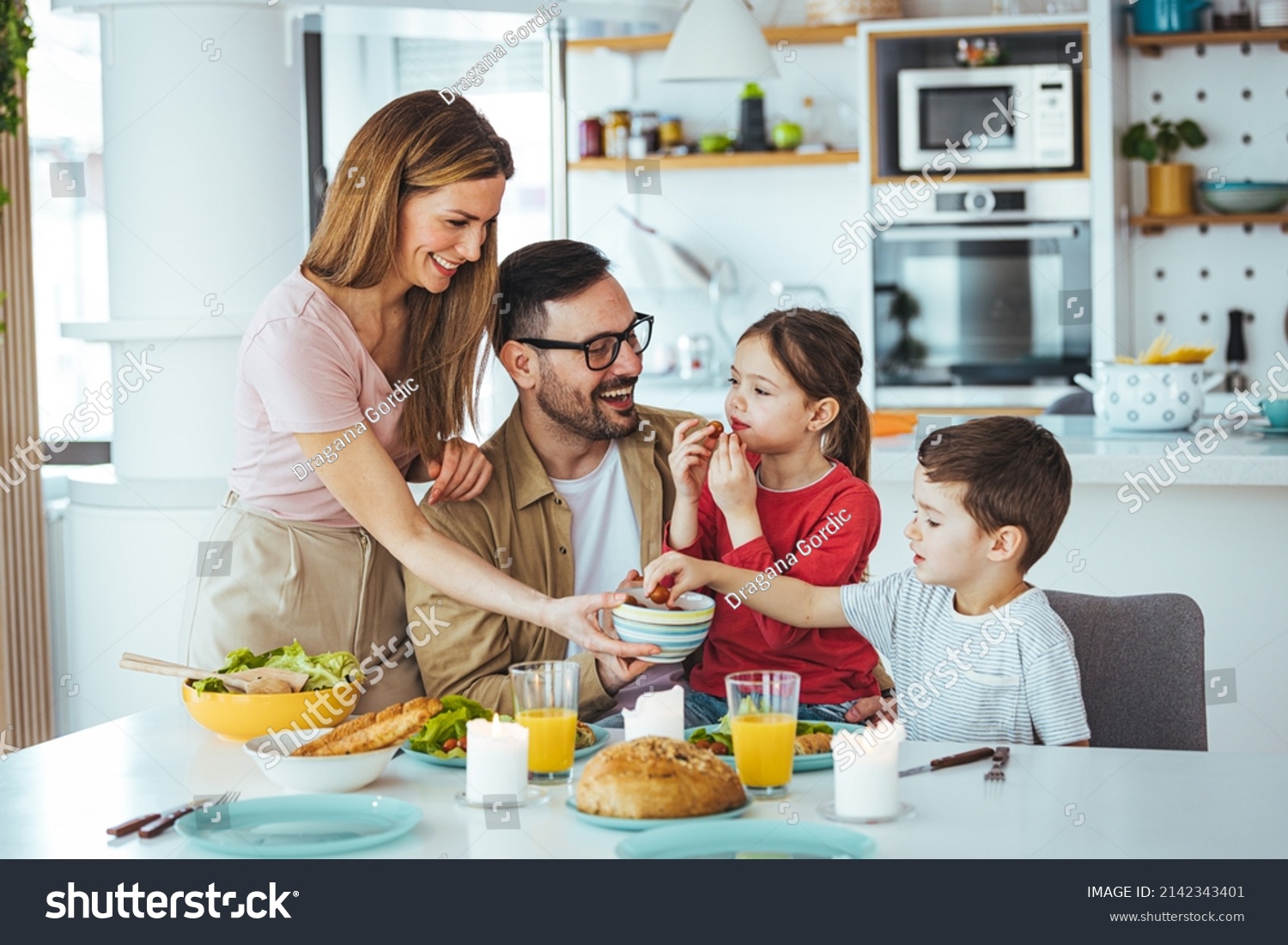 Family of four at the table, mother serves them breakfast. Everyone is happy and smiling, parents enjoy spending time with their children. A cheerful family has fun during a meal at the dining table. #2142343401